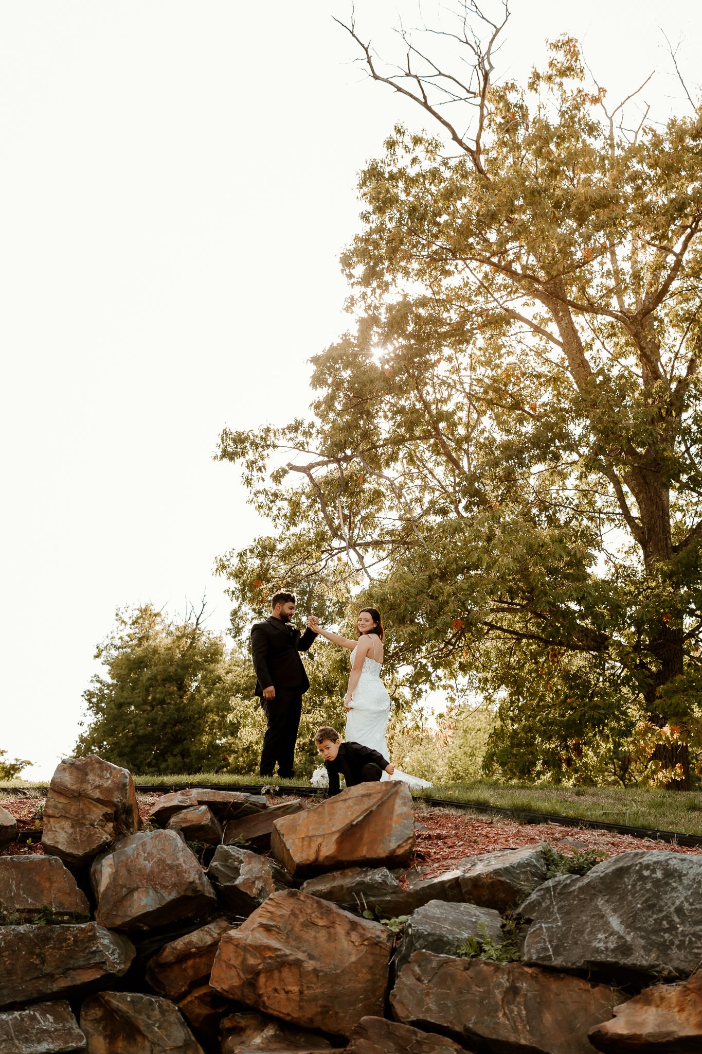 Bride in a white gown and groom in a black suit share a moment atop a stone wall beneath a large tree while a photographer crouches nearby.