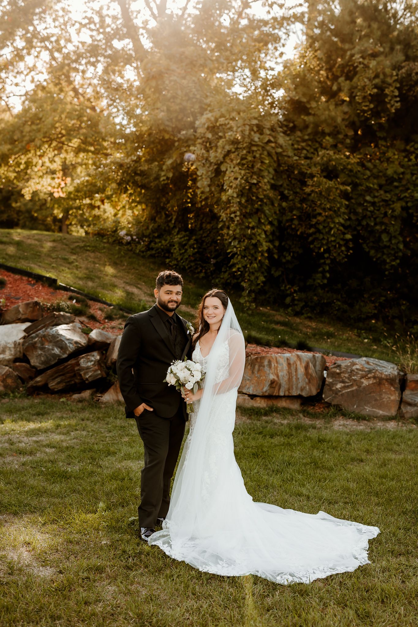 Bride in a white lace gown with a long train and veil holds a bouquet beside her groom in a black suit on a sunlit lawn by a stone wall.