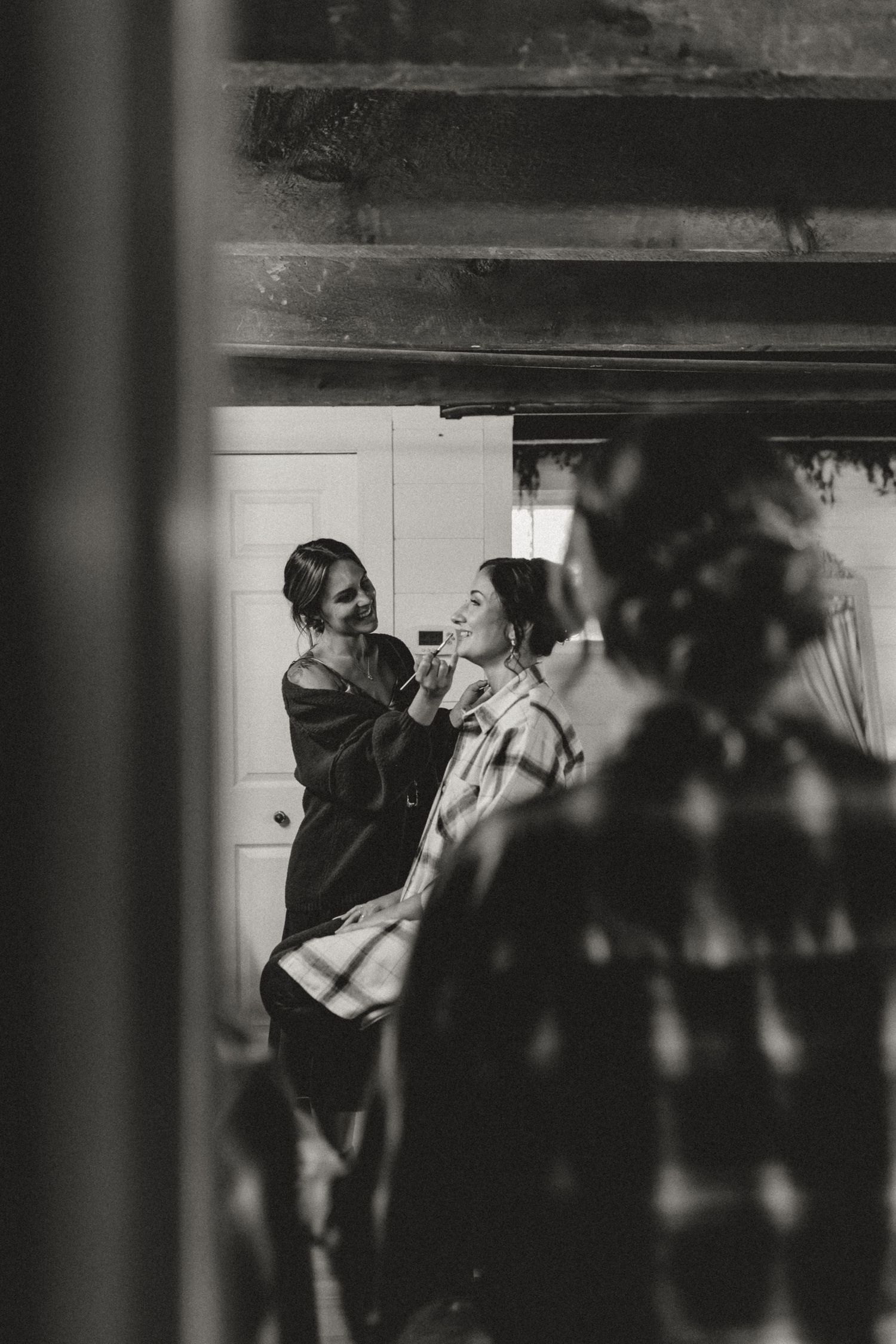 Black and white photo of a makeup artist applying makeup to a woman in a plaid shirt in a rustic room with exposed wooden beams.