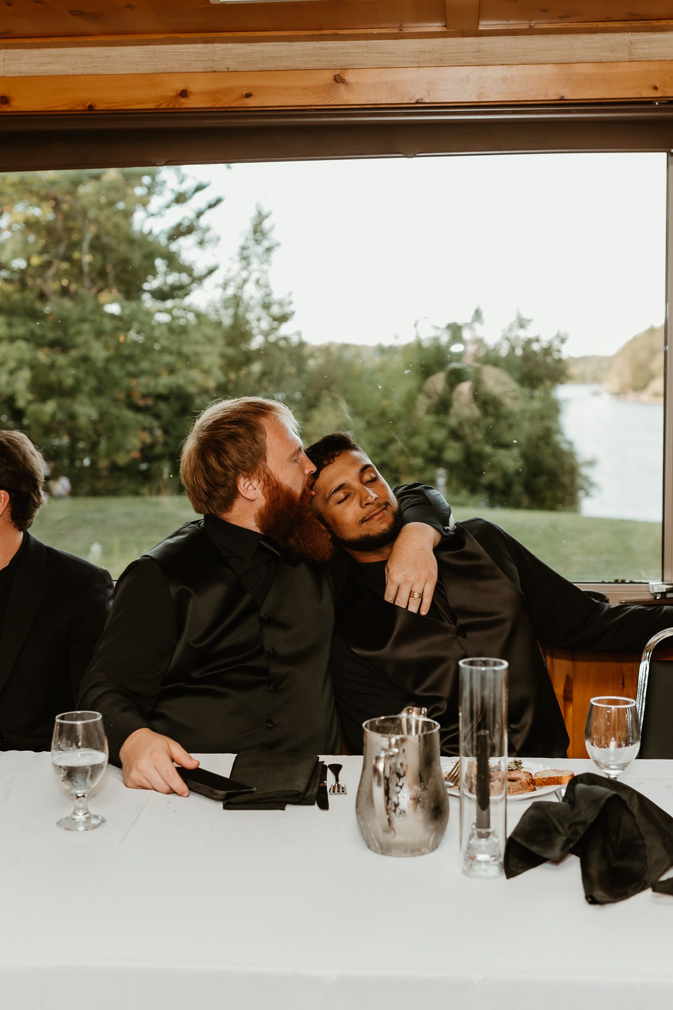 Two men in black shirts embrace affectionately at a white-clothed table near a lakeside window during a wedding reception.