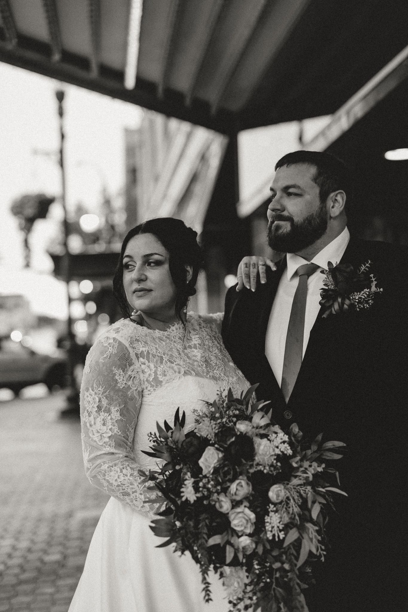 Black and white photo of a bride in a lace gown holding a floral bouquet beside a bearded groom in a suit on a city street.