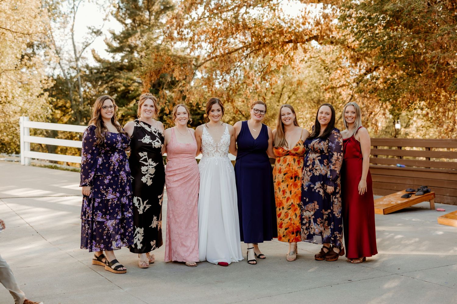 Eight women in colorful dresses, including one in white, pose together outdoors on a patio surrounded by autumn trees.