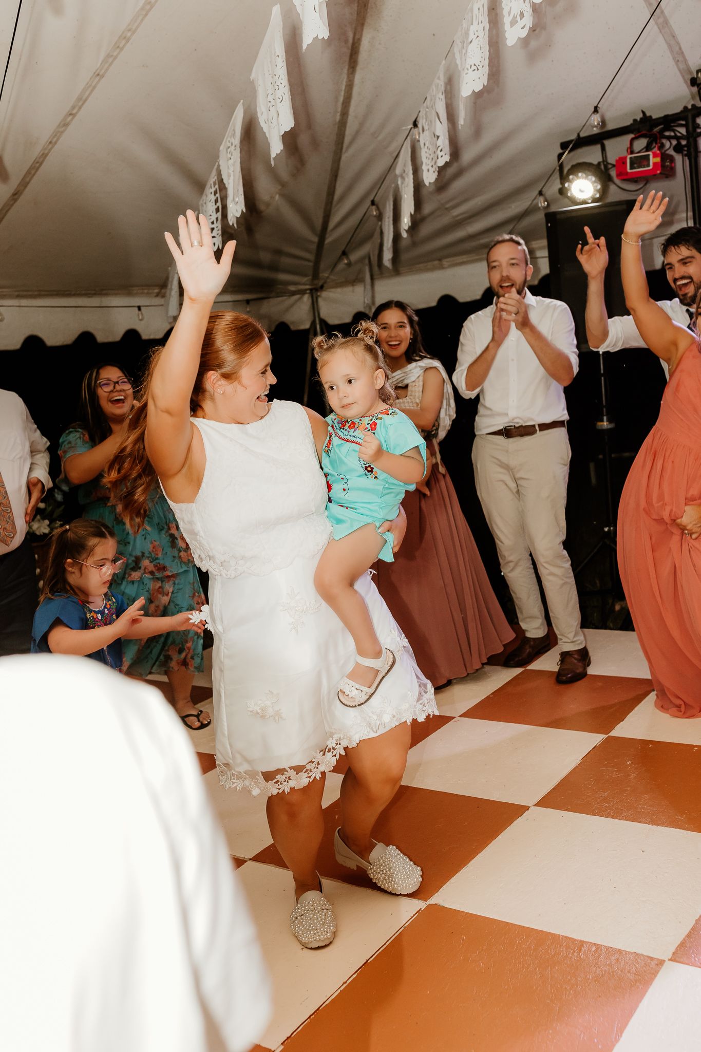 Woman in white dress holding toddler dancing at wedding reception on checkered dance floor under tent.