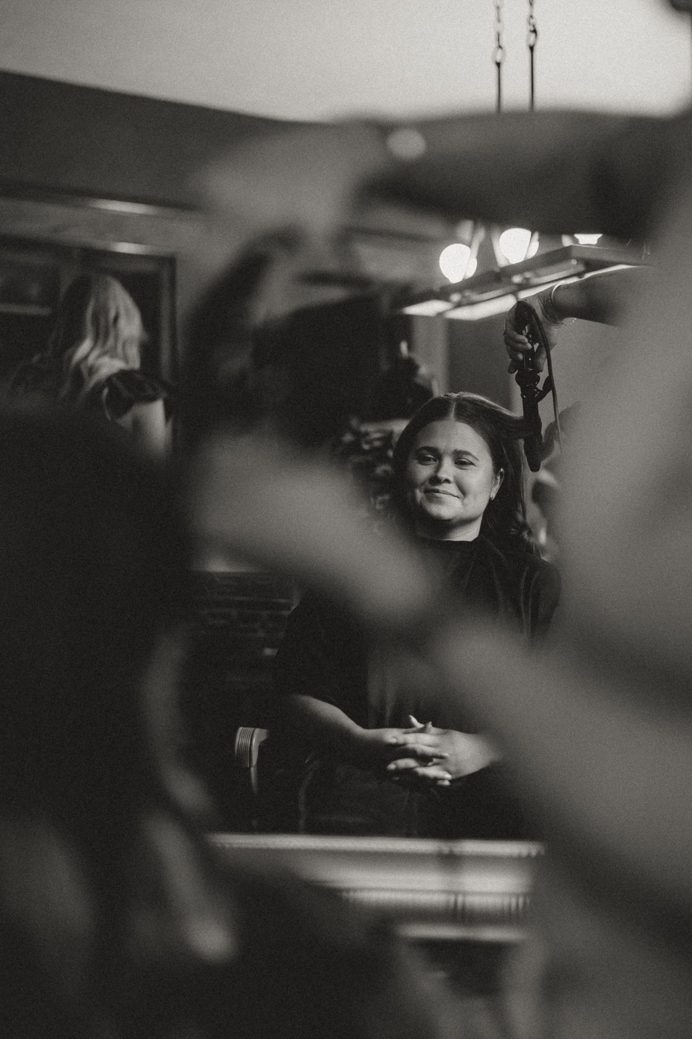 Black and white photo of a smiling woman seated in a salon chair under bright studio lights.