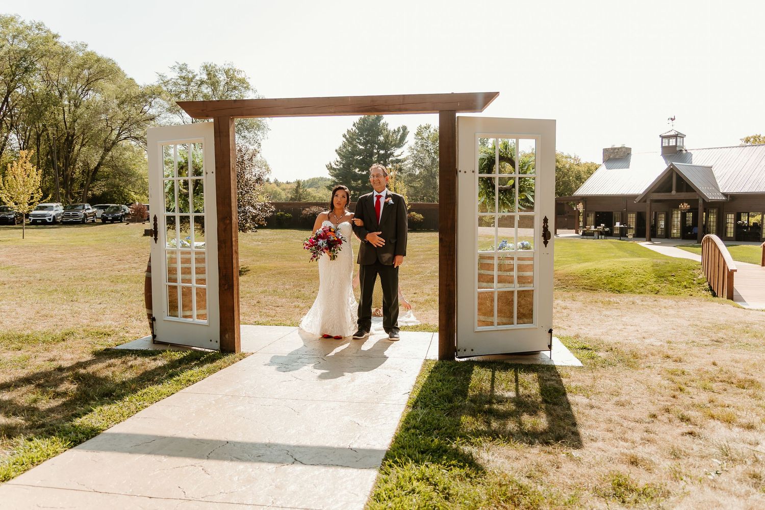 Bride and groom standing under rustic wooden arch with French doors at outdoor wedding ceremony on sunny day.