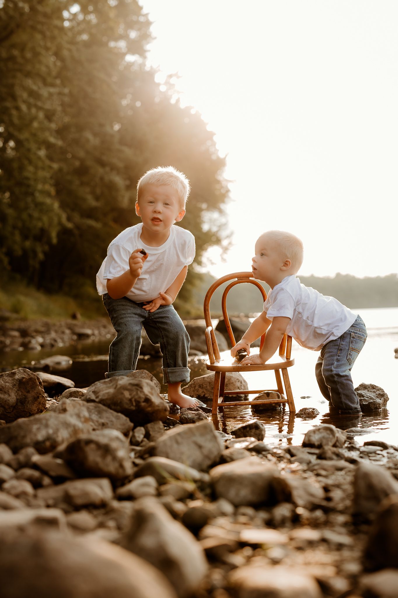 Two young boys playing on rocky shoreline near water at golden hour, wearing white shirts and jeans.