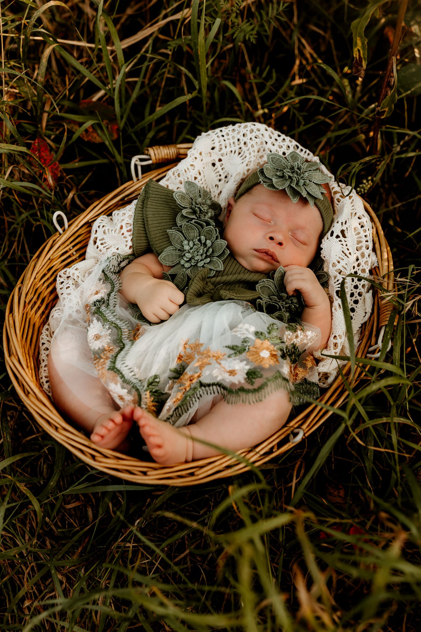 Sleeping newborn baby in green outfit and floral headband nestled in wicker basket surrounded by lush grass.