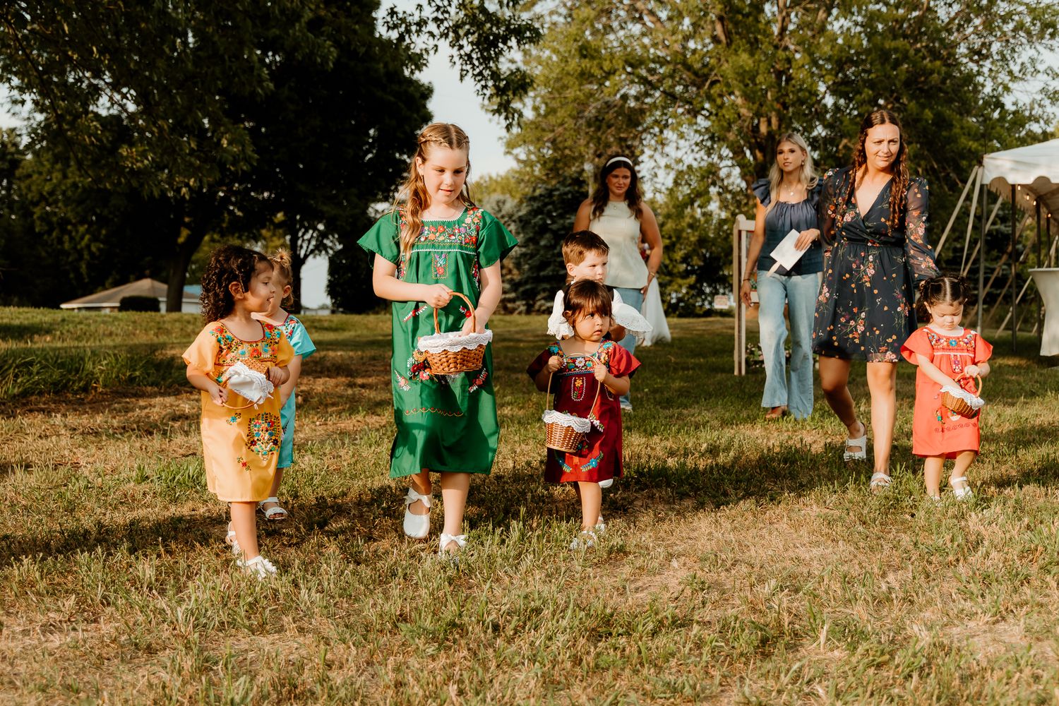 Children and adults enjoy an outdoor gathering on green grass, with girls in colorful dresses playing together.