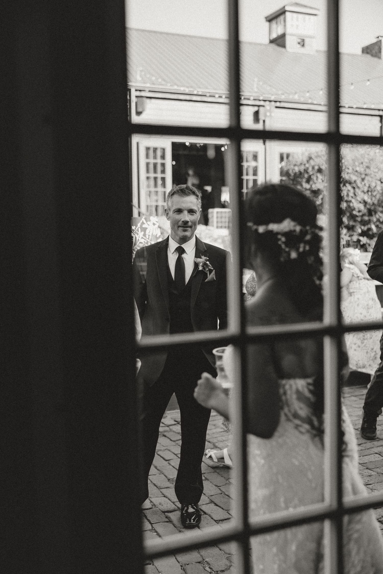 Black and white wedding photo of groom gazing at bride through elegant glass doors at outdoor venue.