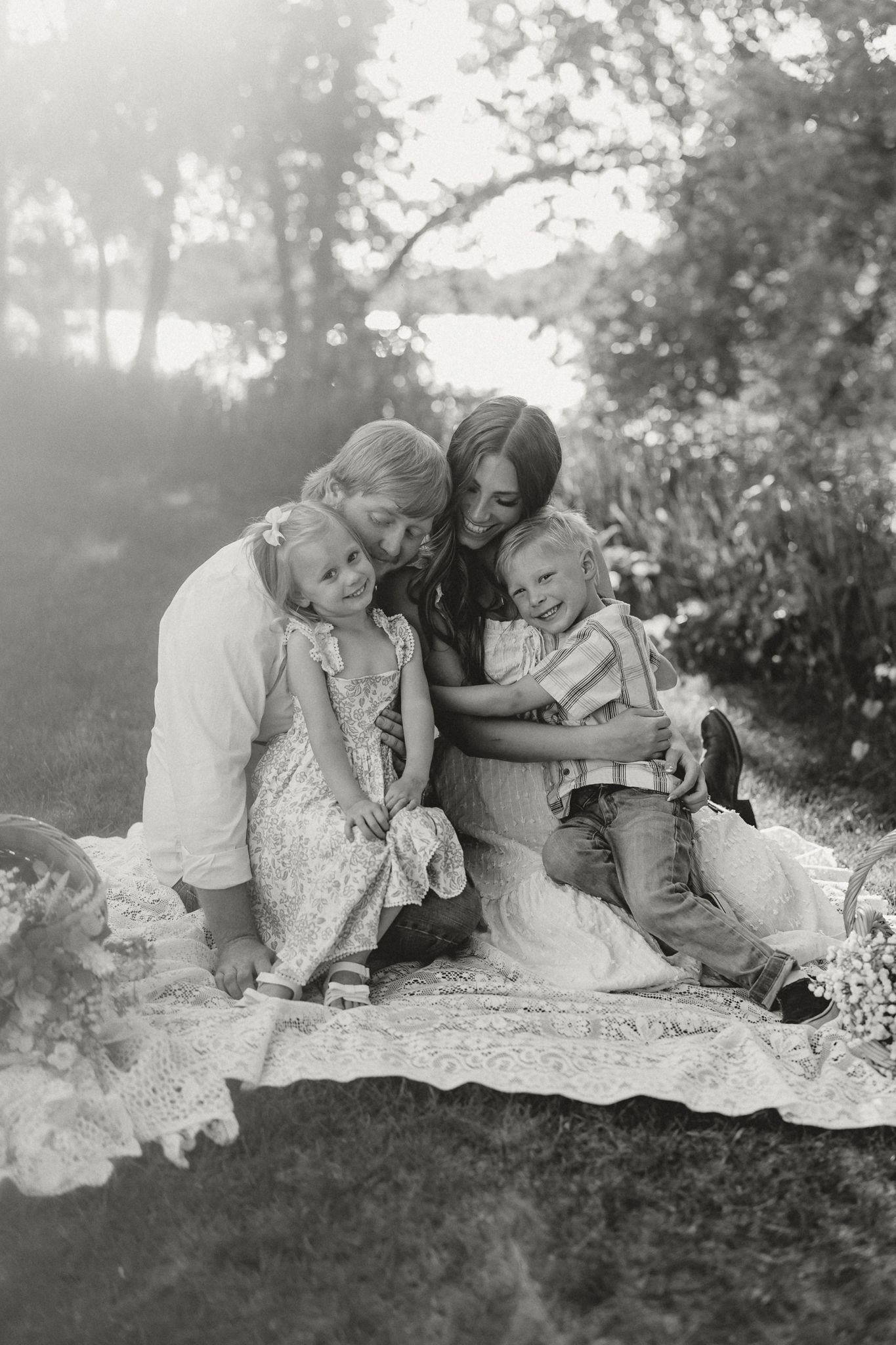 Black and white photo of family of four cuddling together on blanket in sunny park.