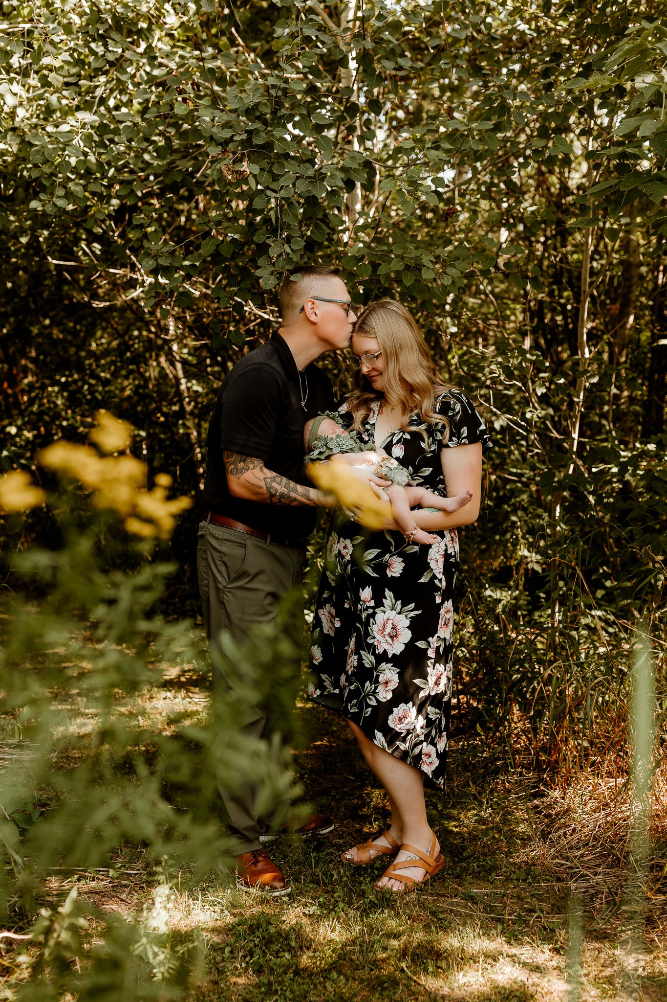 Couple sharing a tender moment in a lush garden, woman in floral dress holding a yellow bouquet, man with tattoos kissing her head.