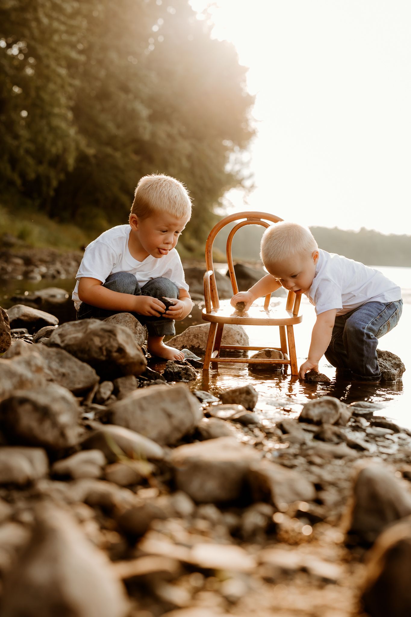 Two young boys playing with rocks and a wooden chair by a sunlit rocky riverbank surrounded by trees.