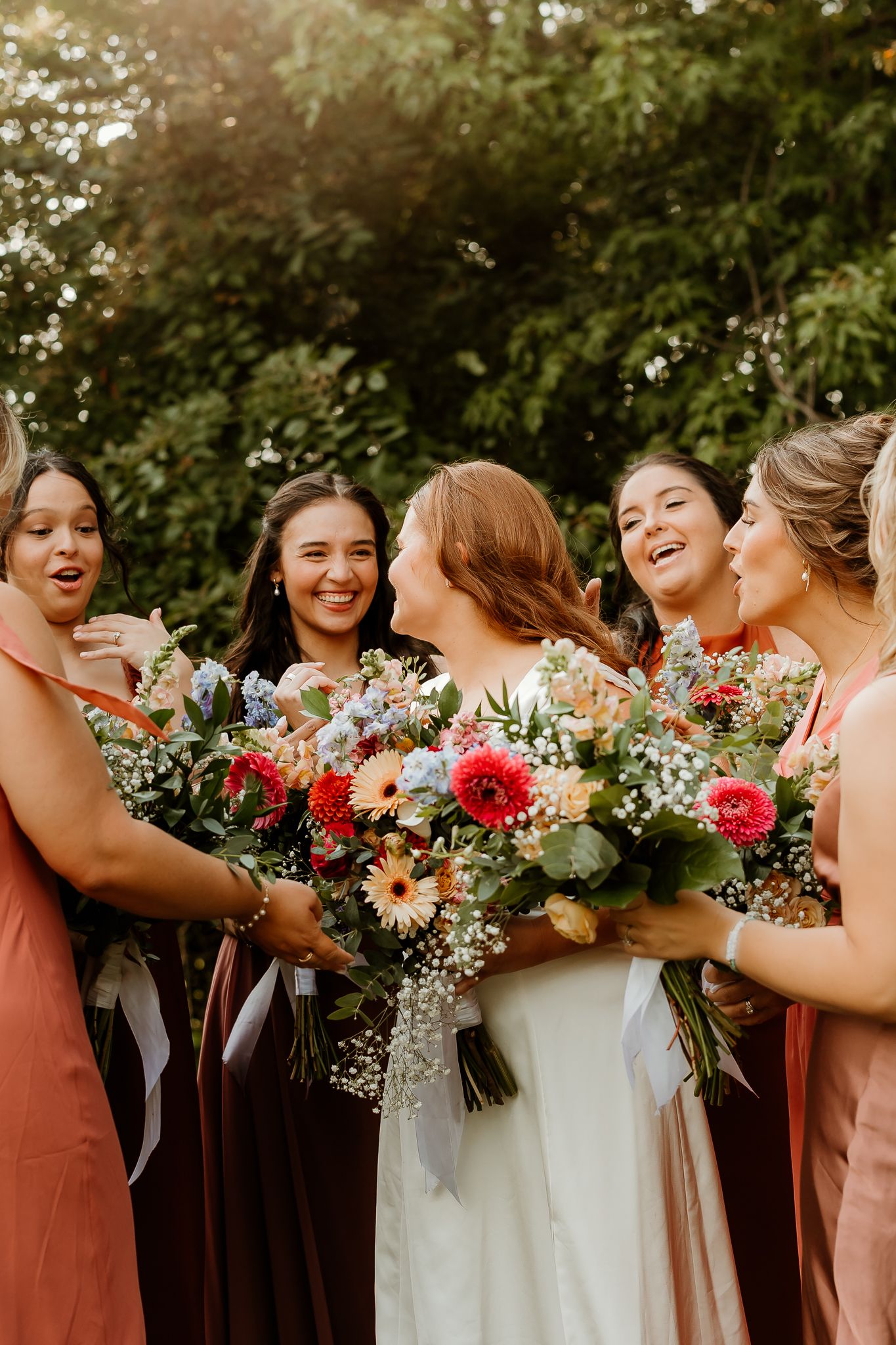 Bride and bridesmaids in coral dresses laughing together while holding colorful wildflower bouquets outdoors.