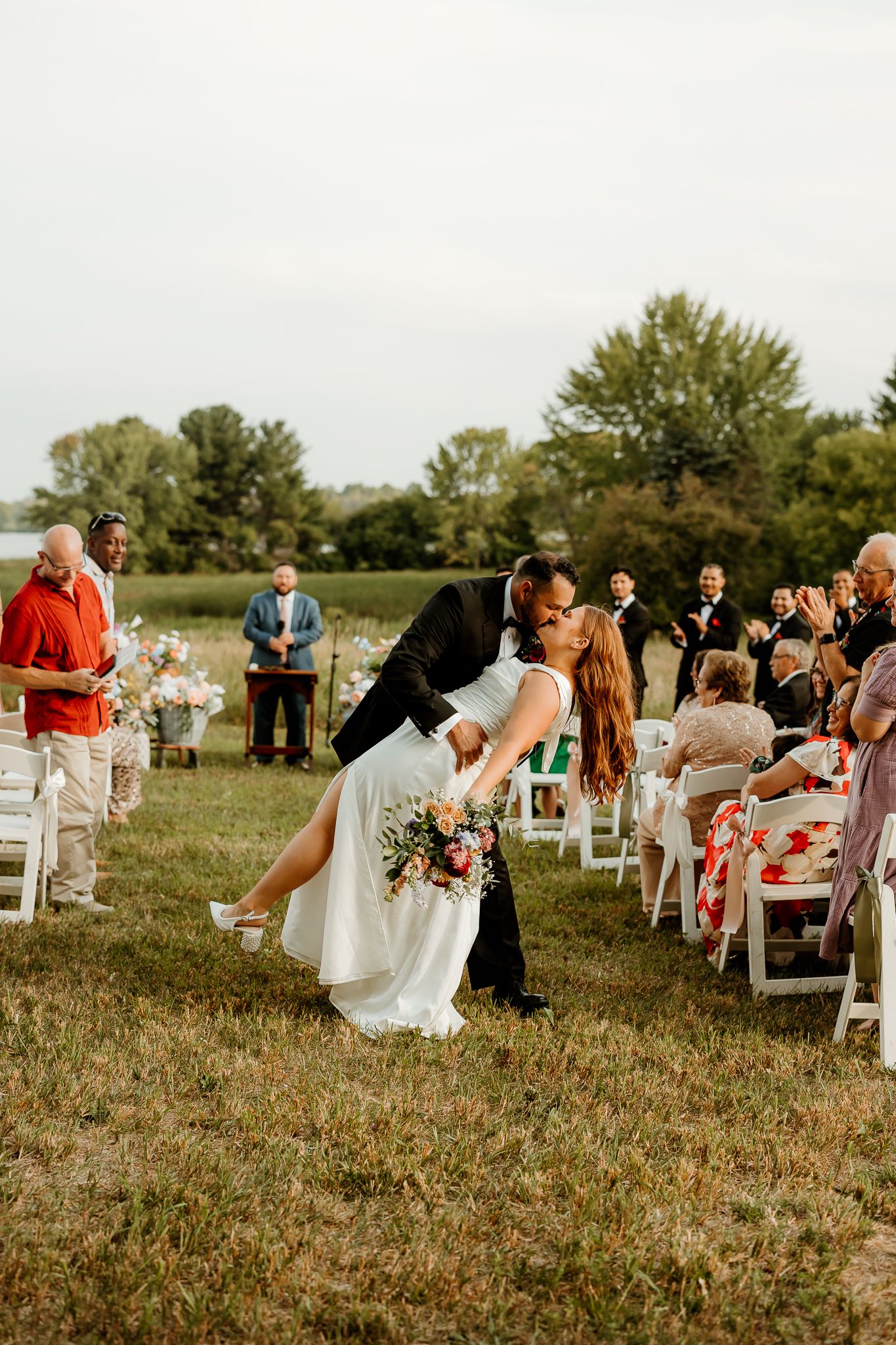 A groom dips his bride for a kiss during an outdoor wedding ceremony while guests cheer along a white chair-lined aisle.