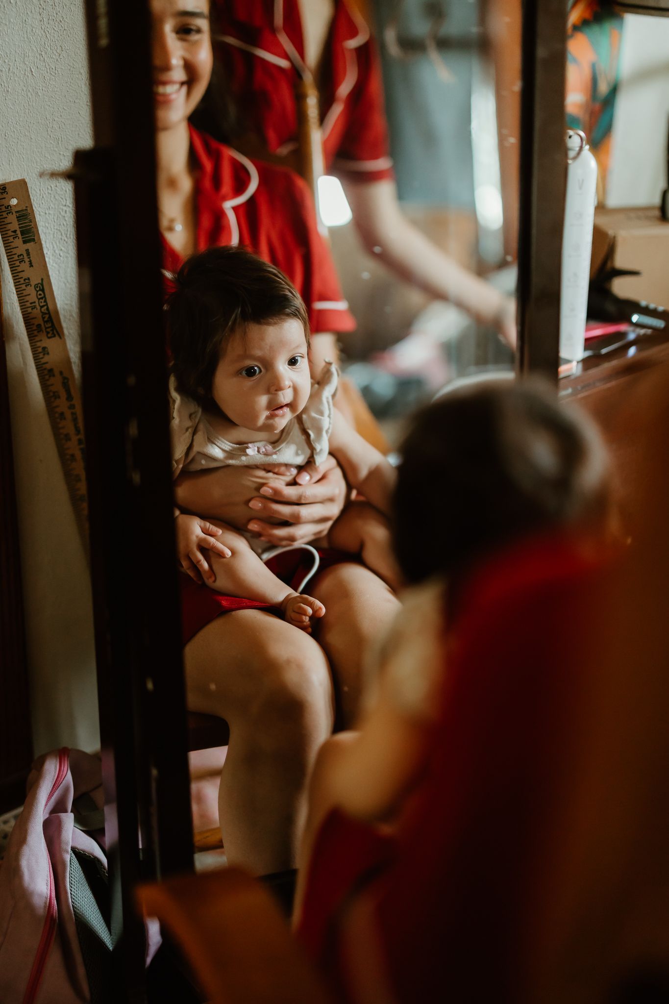 A smiling woman in red holds a cute baby on her lap, reflected in a mirror in a cozy indoor setting.