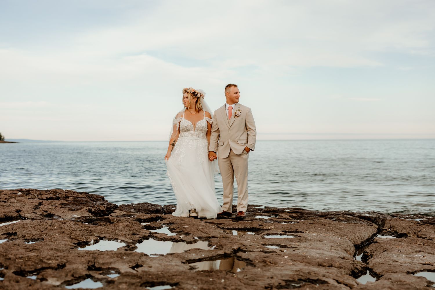 Bride and groom holding hands walking on rocky shoreline with ocean backdrop on their wedding day.