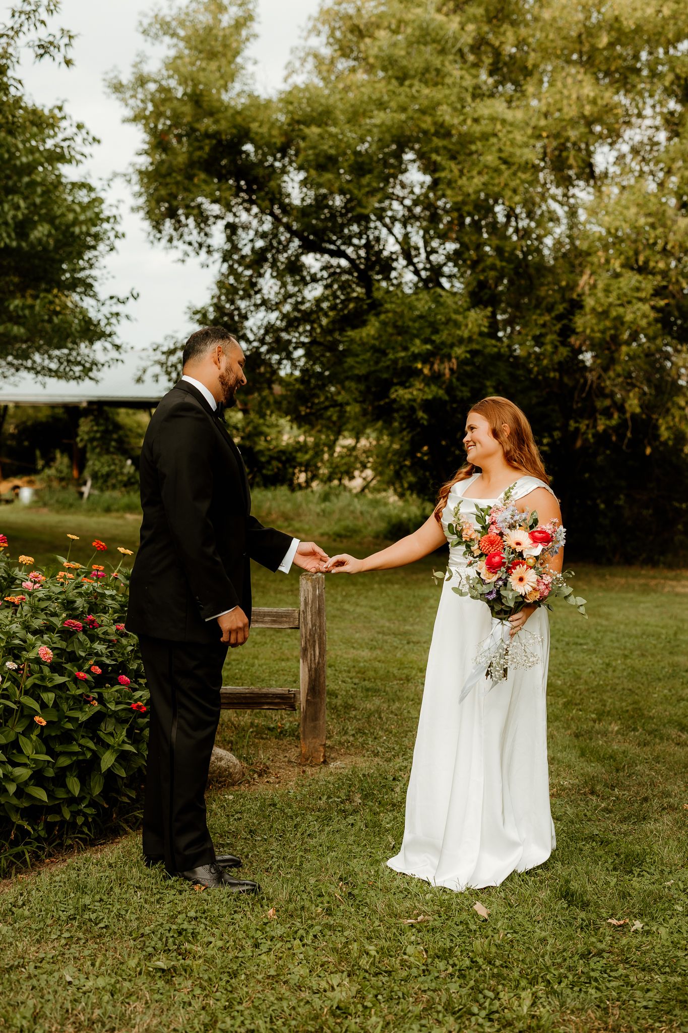 Bride and groom hold hands outdoors on green lawn, she holds colorful bouquet, surrounded by lush trees and flowers.