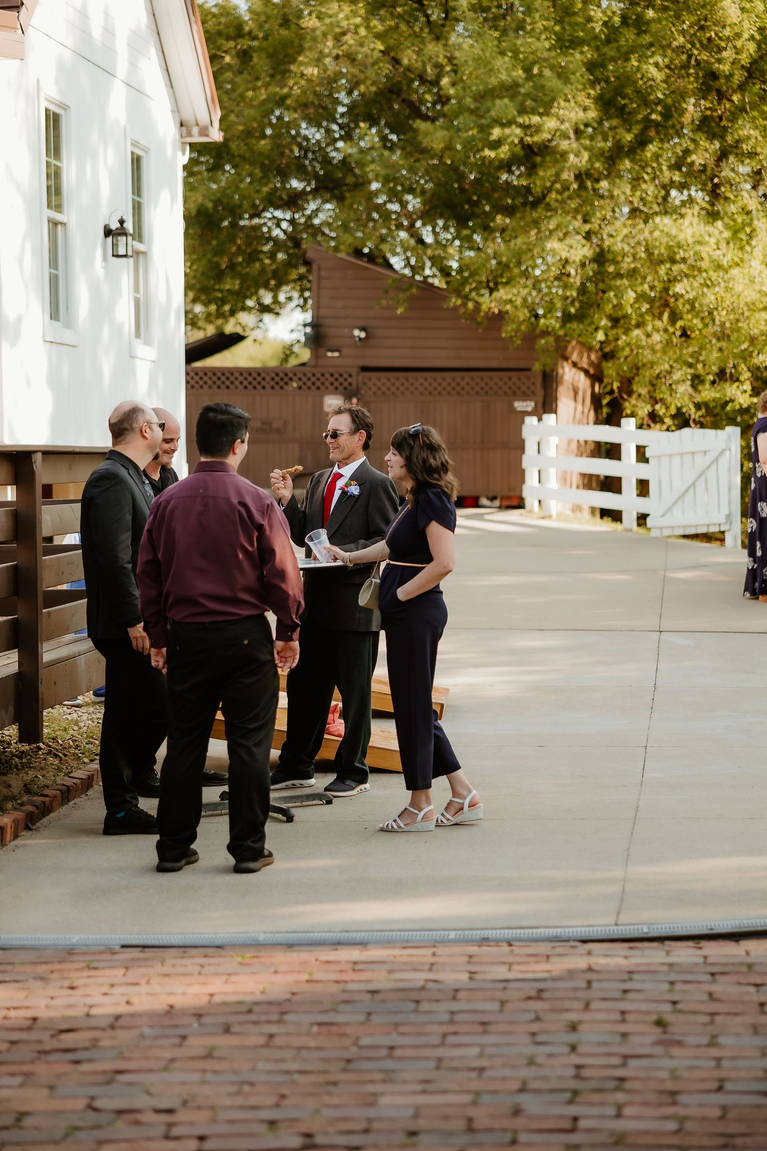 Group of people conversing outdoors near a white barn venue on a sunny day at a wedding event.