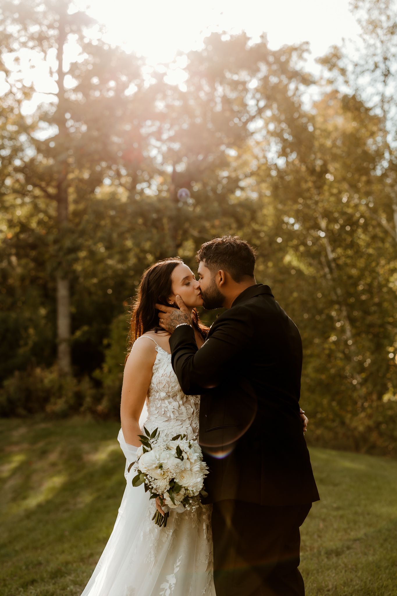Bride in a lace wedding gown holding a white bouquet shares a kiss with her groom in a dark suit on a sunlit grassy lawn.