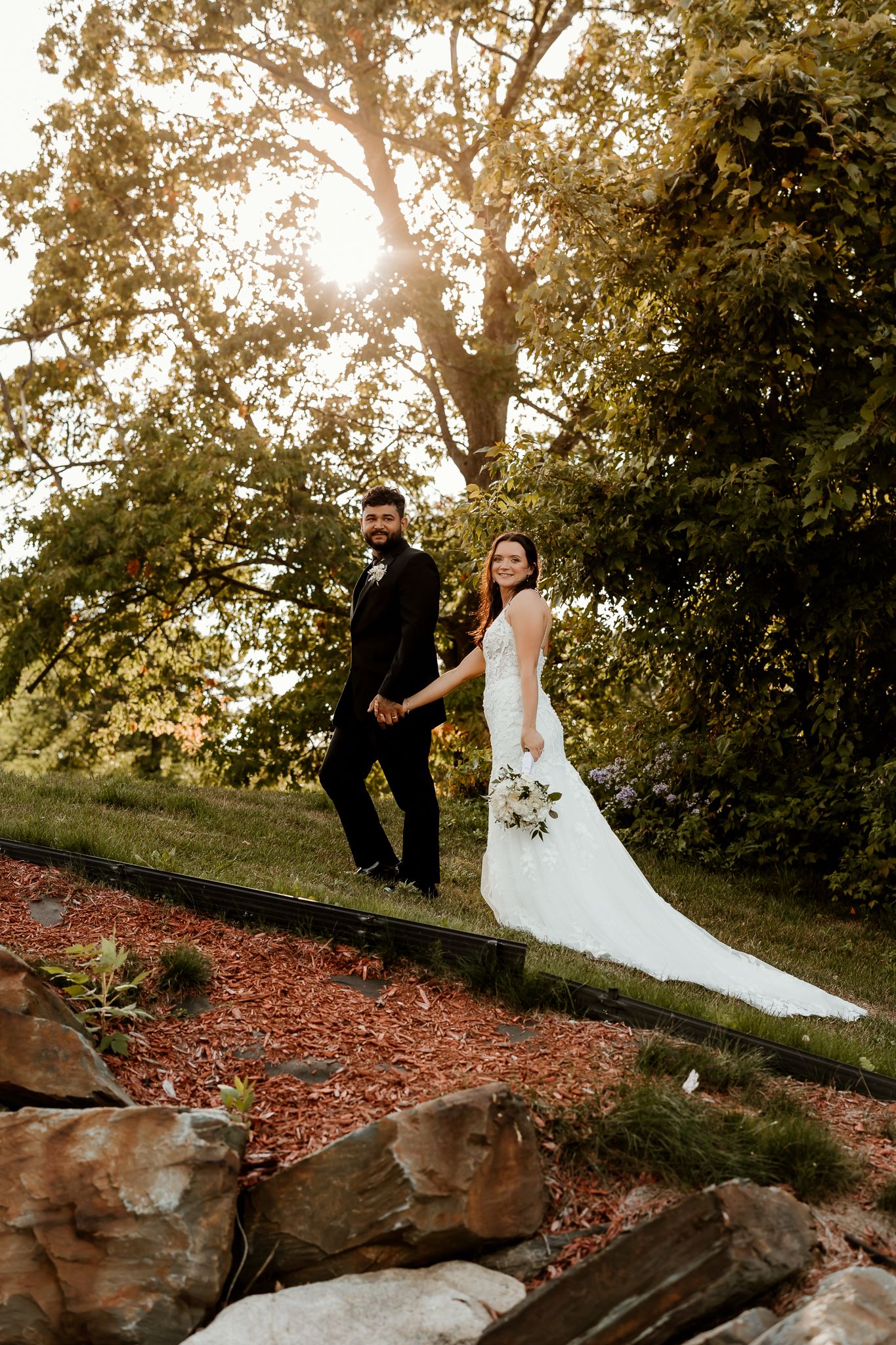 Bride in a white lace wedding gown and groom in a dark suit hold hands while walking up a landscaped garden path in golden sunset light.