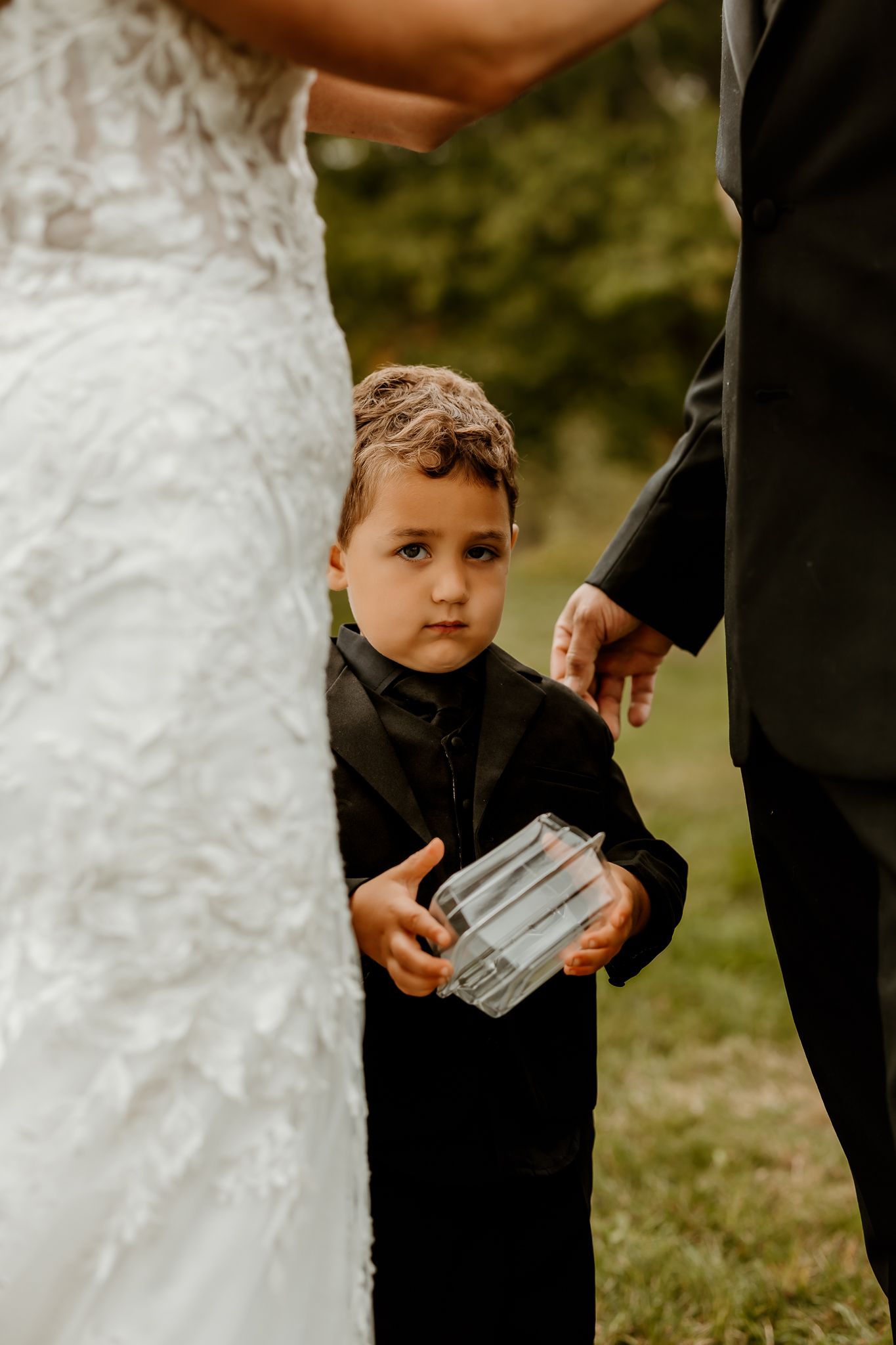 Young ring bearer in a black suit holds a clear box beside a bride in a lace gown during an outdoor wedding ceremony.