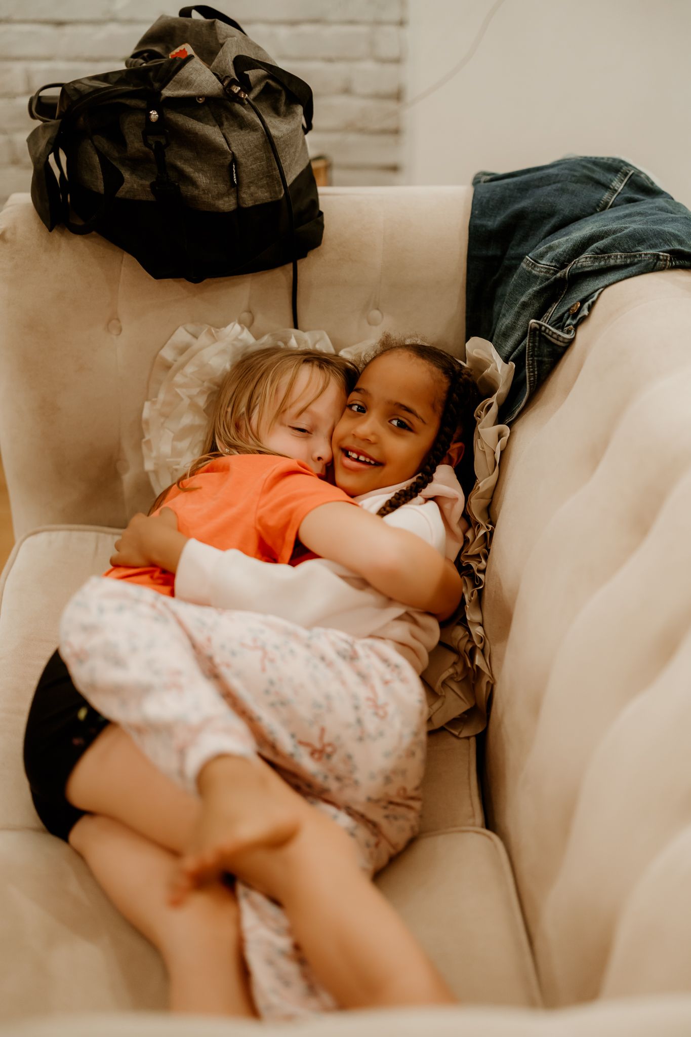 Two young girls hug and smile while cuddling on a cream tufted sofa, with a black duffel bag and denim jacket nearby.