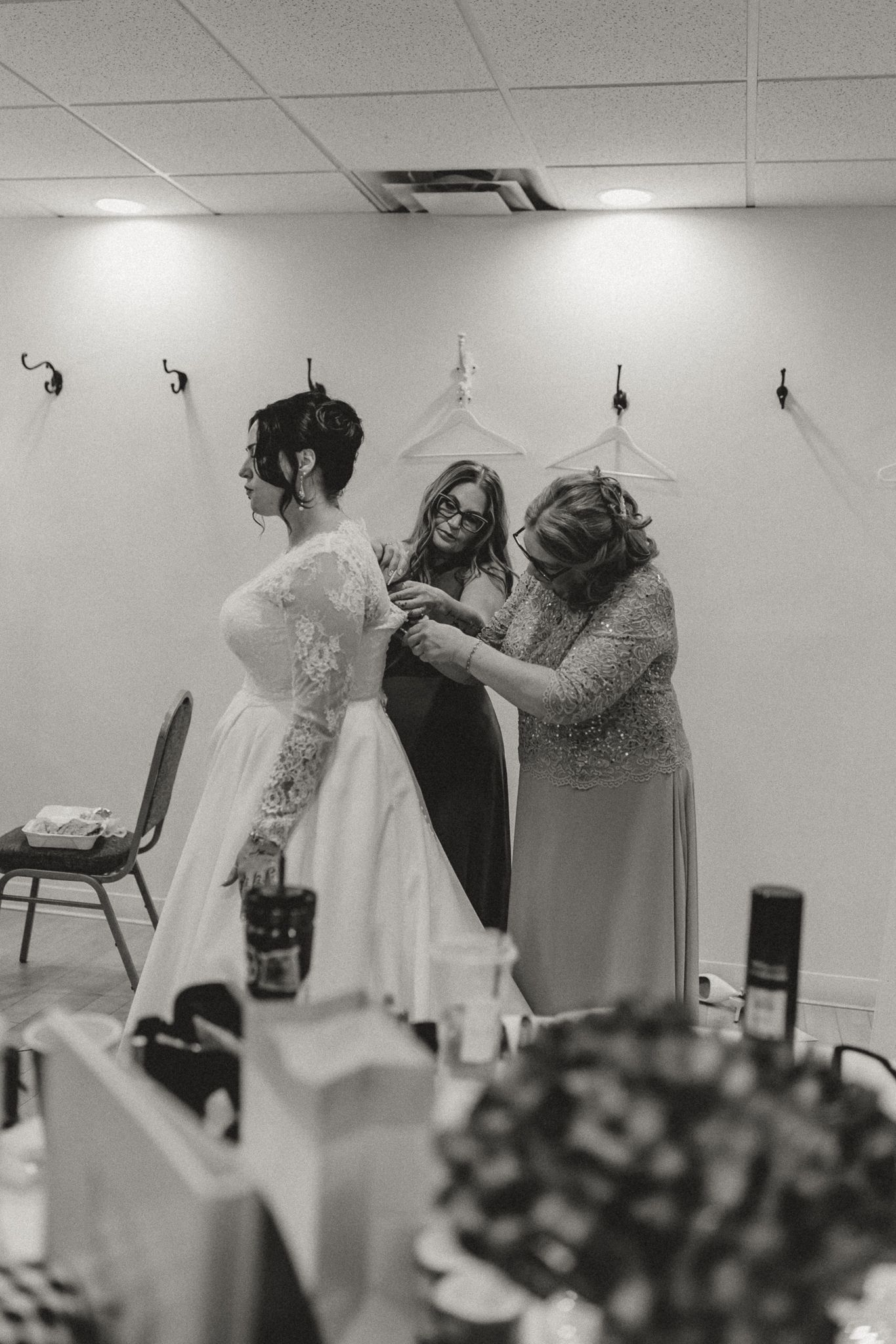 Two women help a bride in a lace wedding gown get ready in a bright room in this black and white photo.