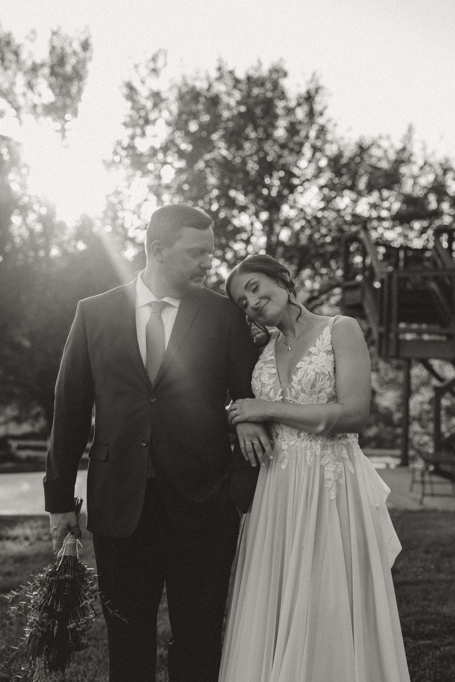 Black and white wedding portrait of a bride leaning her head on the groom's shoulder outdoors, with sunlight streaming through the trees.