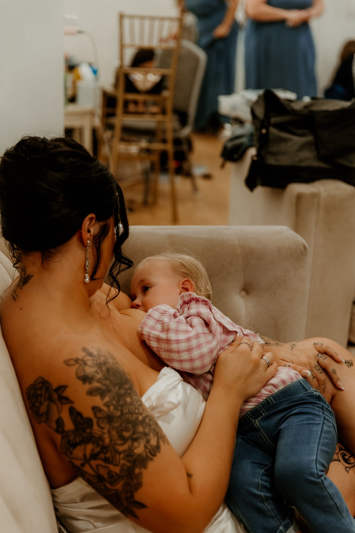A mother with a floral shoulder tattoo and drop earrings nurses her baby in a pink gingham shirt on a cream sofa.
