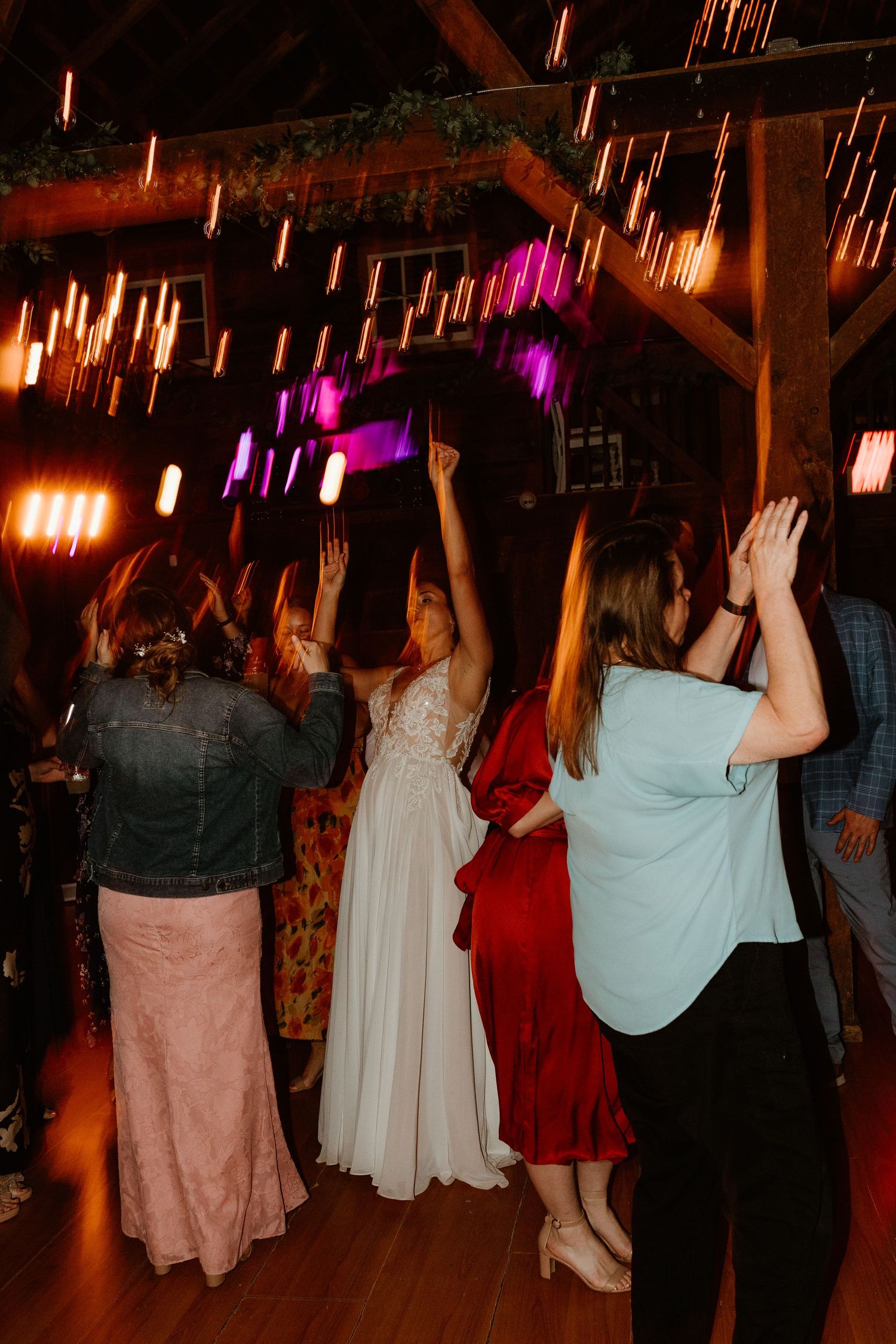 Wedding guests dance with raised hands on a wooden floor in a rustic barn venue lit by string lights and pink and purple uplighting.