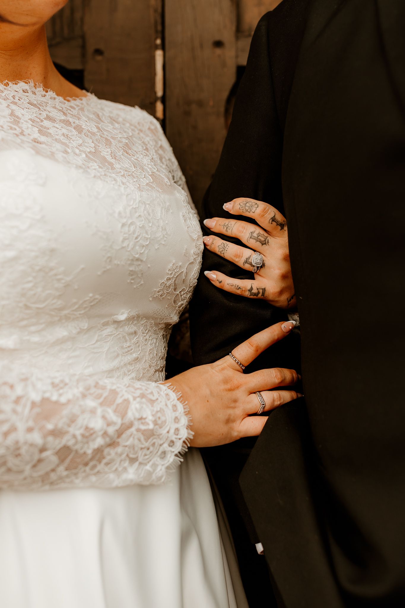 Bride in a white long-sleeved lace wedding dress stands beside a groom in a black suit, his tattooed hand resting on her waist.
