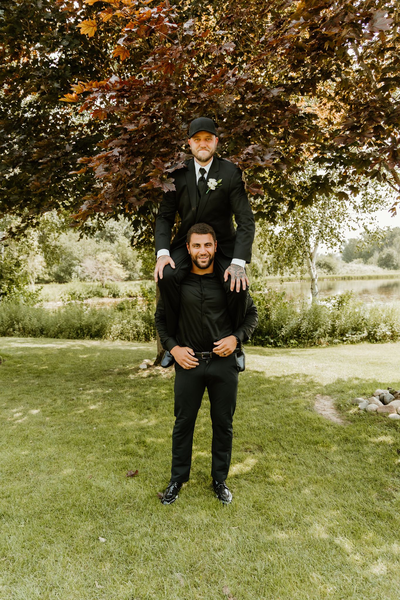 Two men in black suits pose outdoors, one sitting on the other's shoulders, with lush green trees in the background.