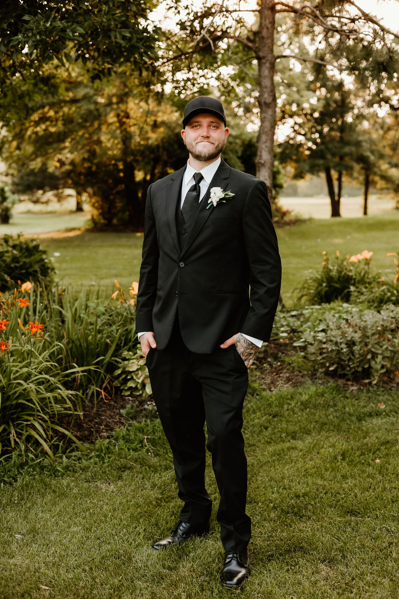 A groom in a black suit, tie, boutonniere, and cap poses outdoors in a lush garden setting.