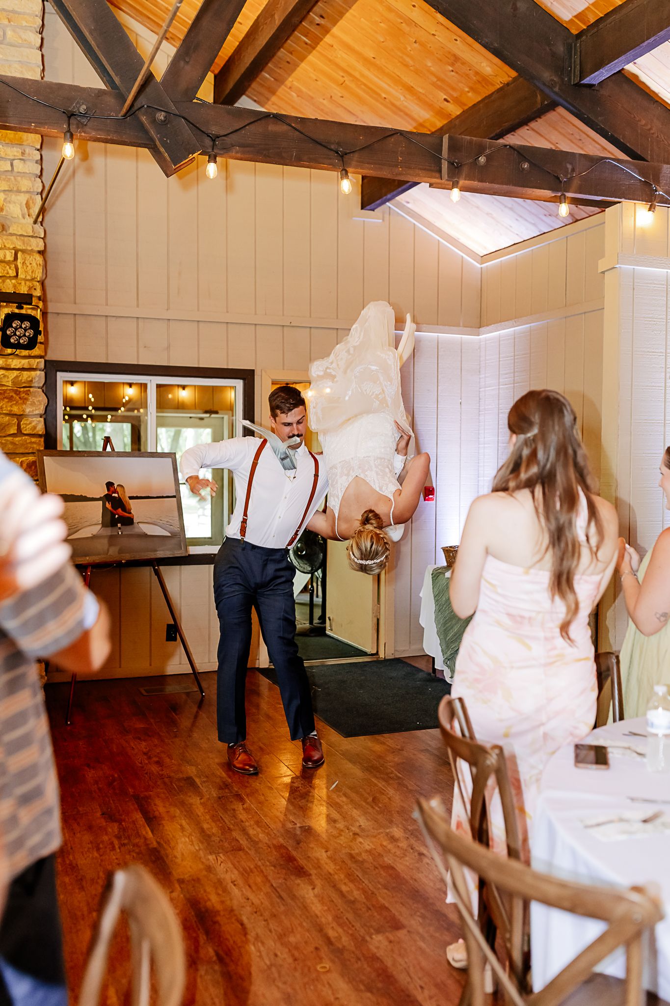 Groom in suspenders lifts bride in wedding dress during lively reception dance in rustic venue with wood beam ceiling.