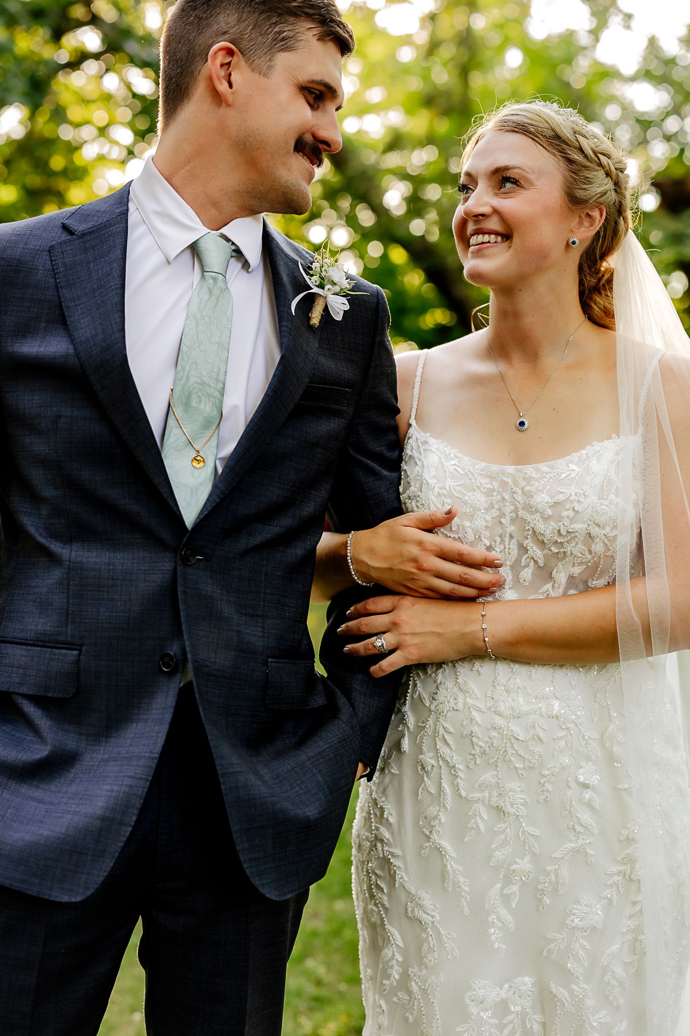 Smiling bride in lace gown and veil stands with groom in navy suit and sage tie outdoors.