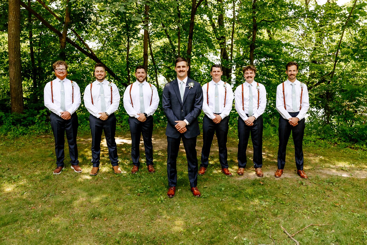 Groom stands centered with seven groomsmen wearing black suits and rust suspenders outdoors in a lush green wooded setting.