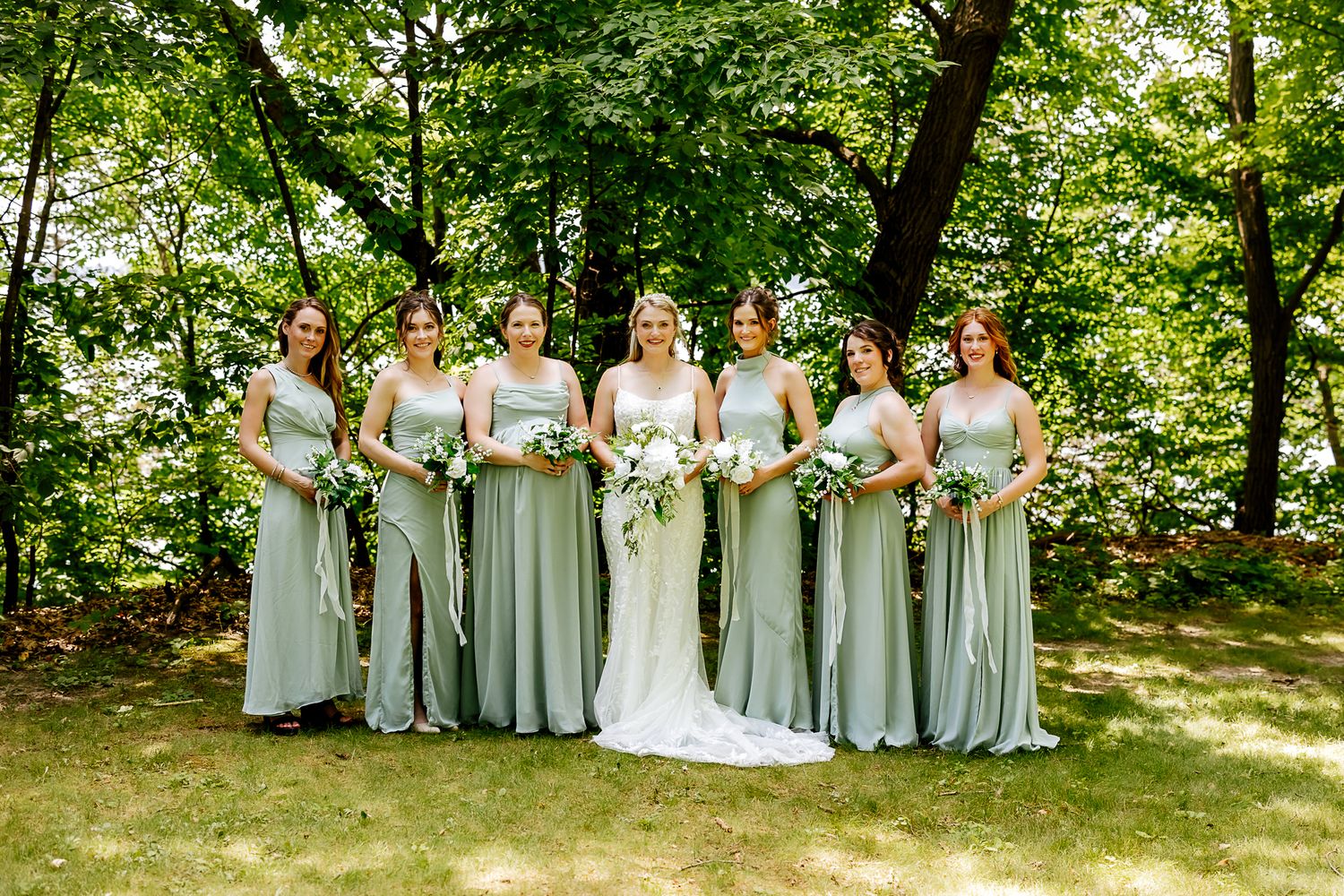 Bride in white gown with five bridesmaids in sage green dresses holding bouquets outdoors in lush green setting.