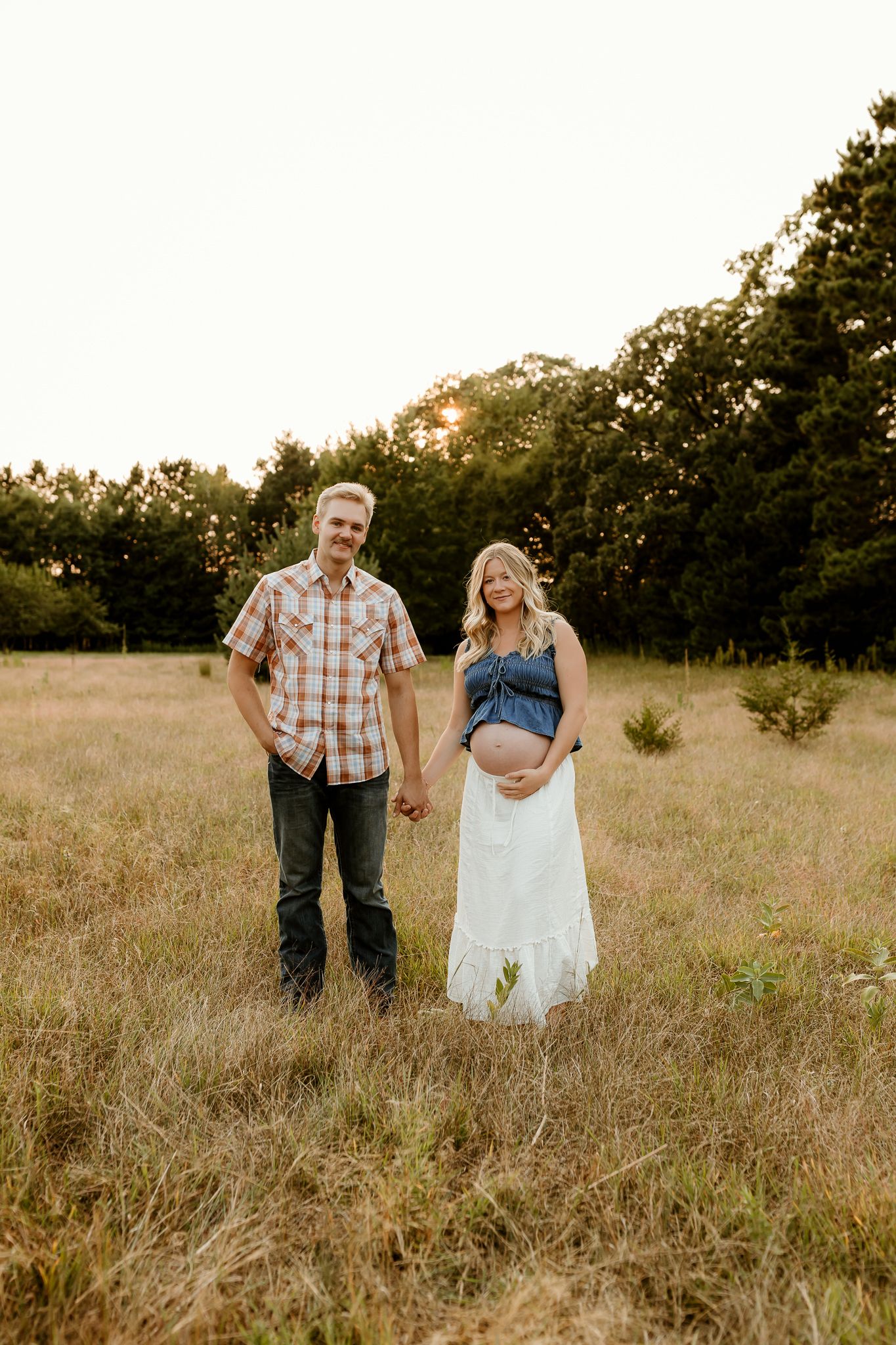 Pregnant couple holding hands in golden field at sunset, woman in white skirt and denim top cradling baby bump.
