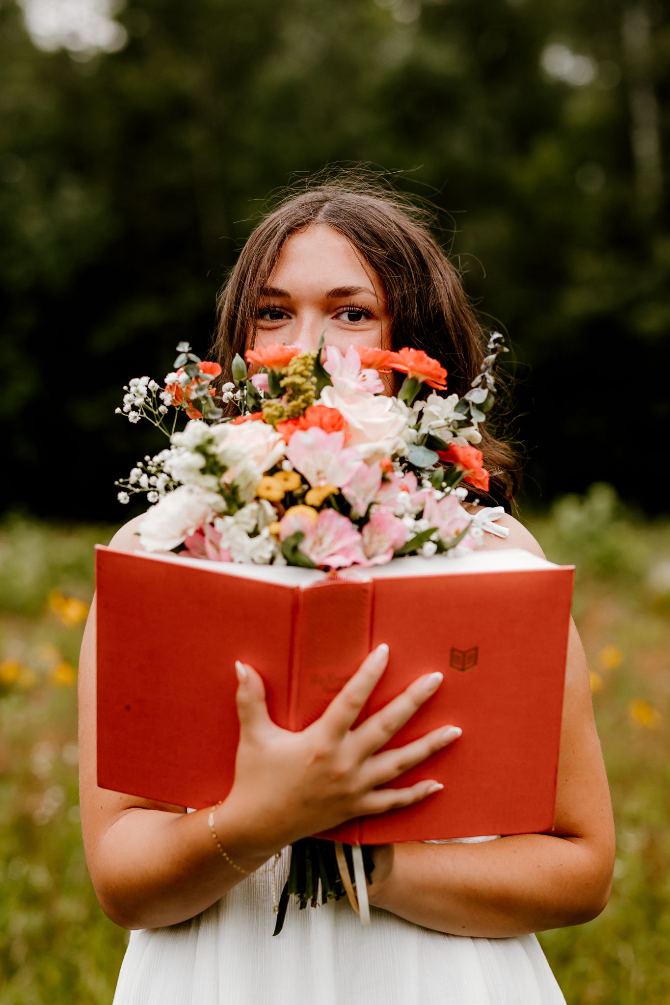 Young woman in white dress holds red book with colorful floral bouquet outdoors in a green meadow.