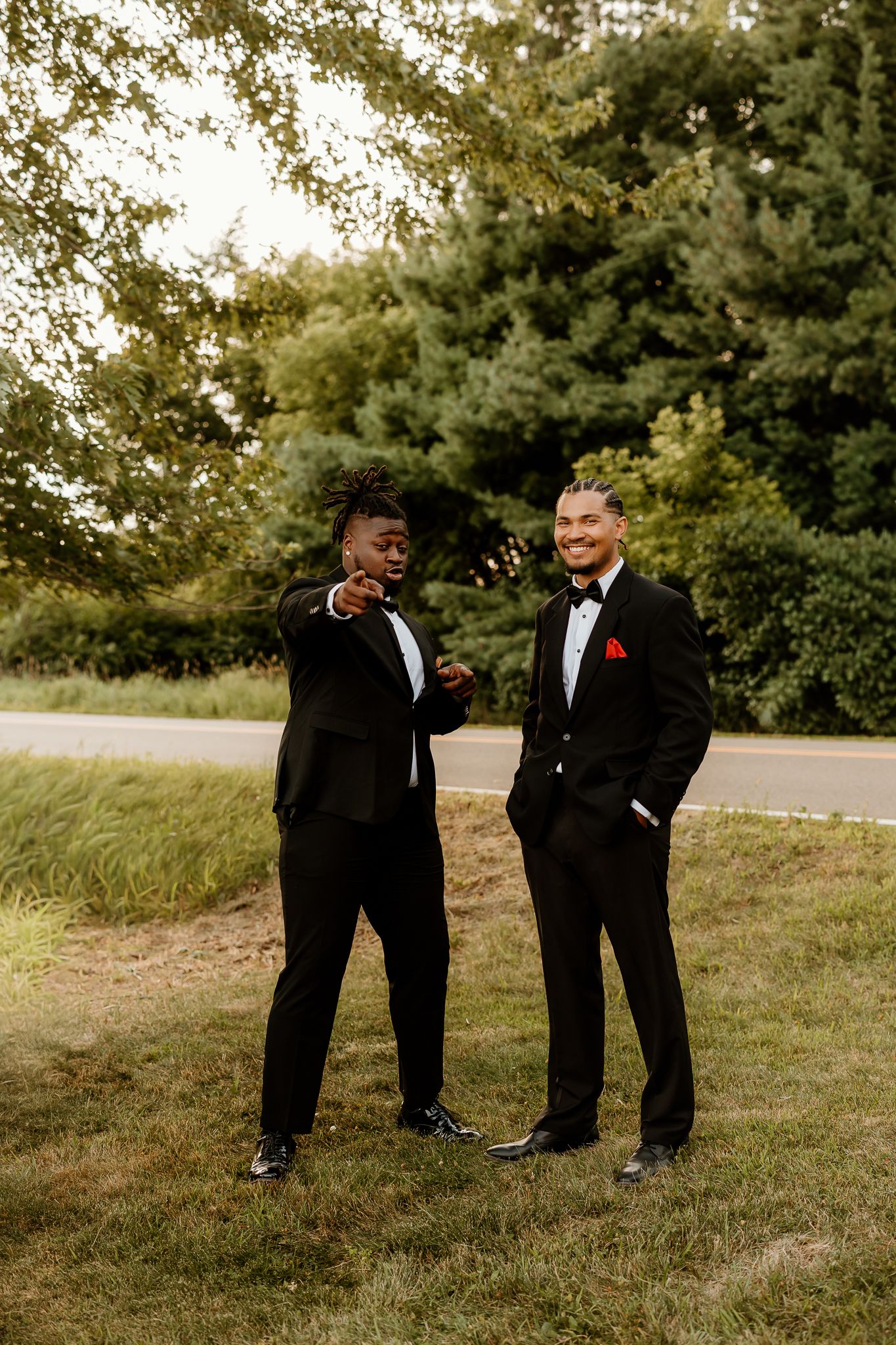 Two men in black tuxedos with bow ties stand outdoors on grass, one pointing and smiling, trees in background.