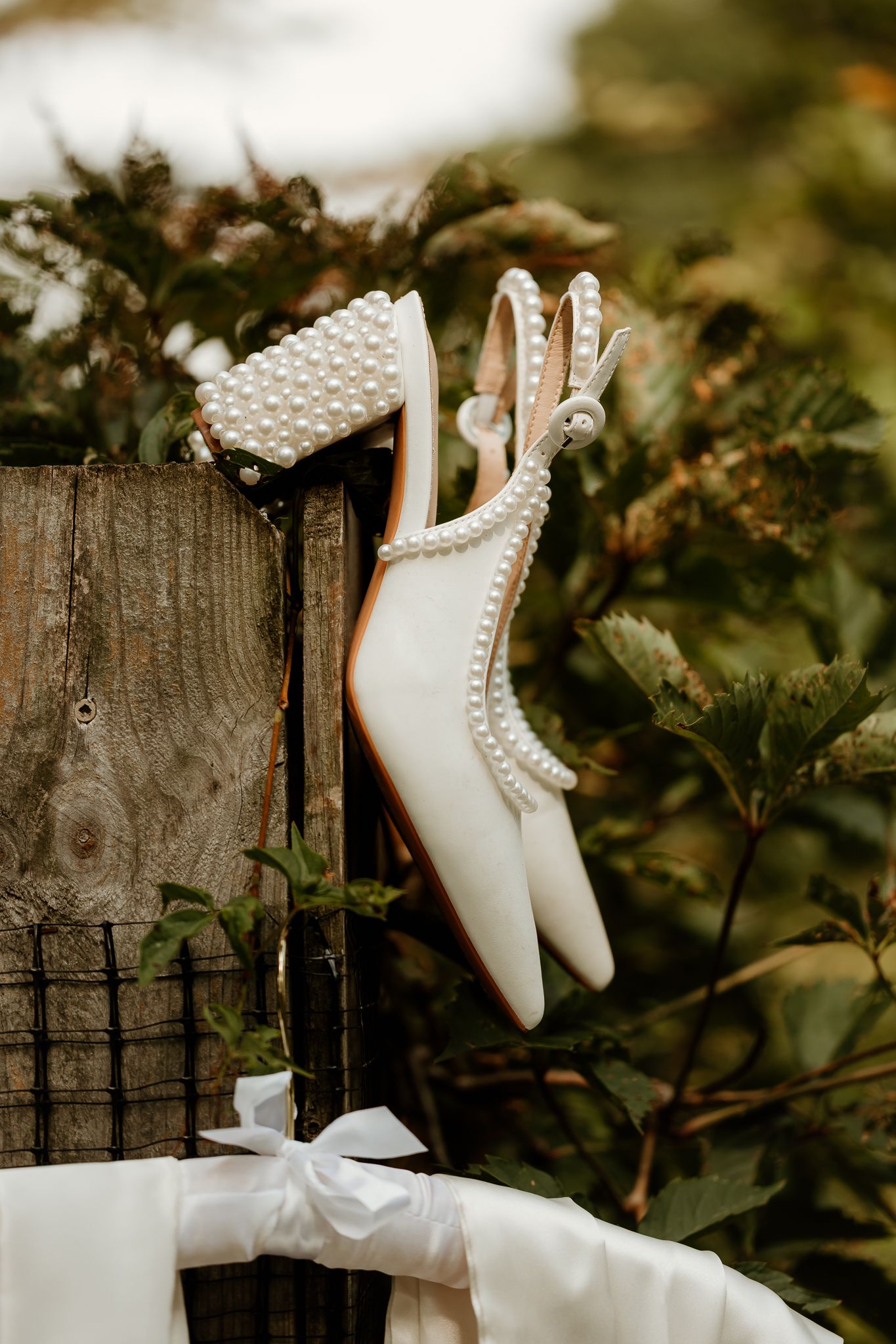 White pearl-embellished bridal heels displayed on a rustic wooden fence surrounded by lush greenery.