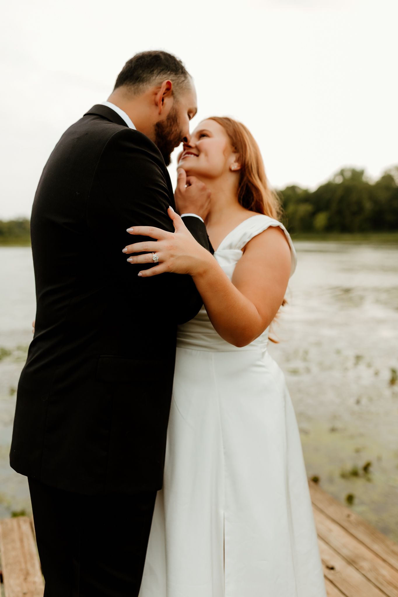 Bride and groom share a romantic moment on a dock by a lake, with the bride in a white gown and groom in a black suit.