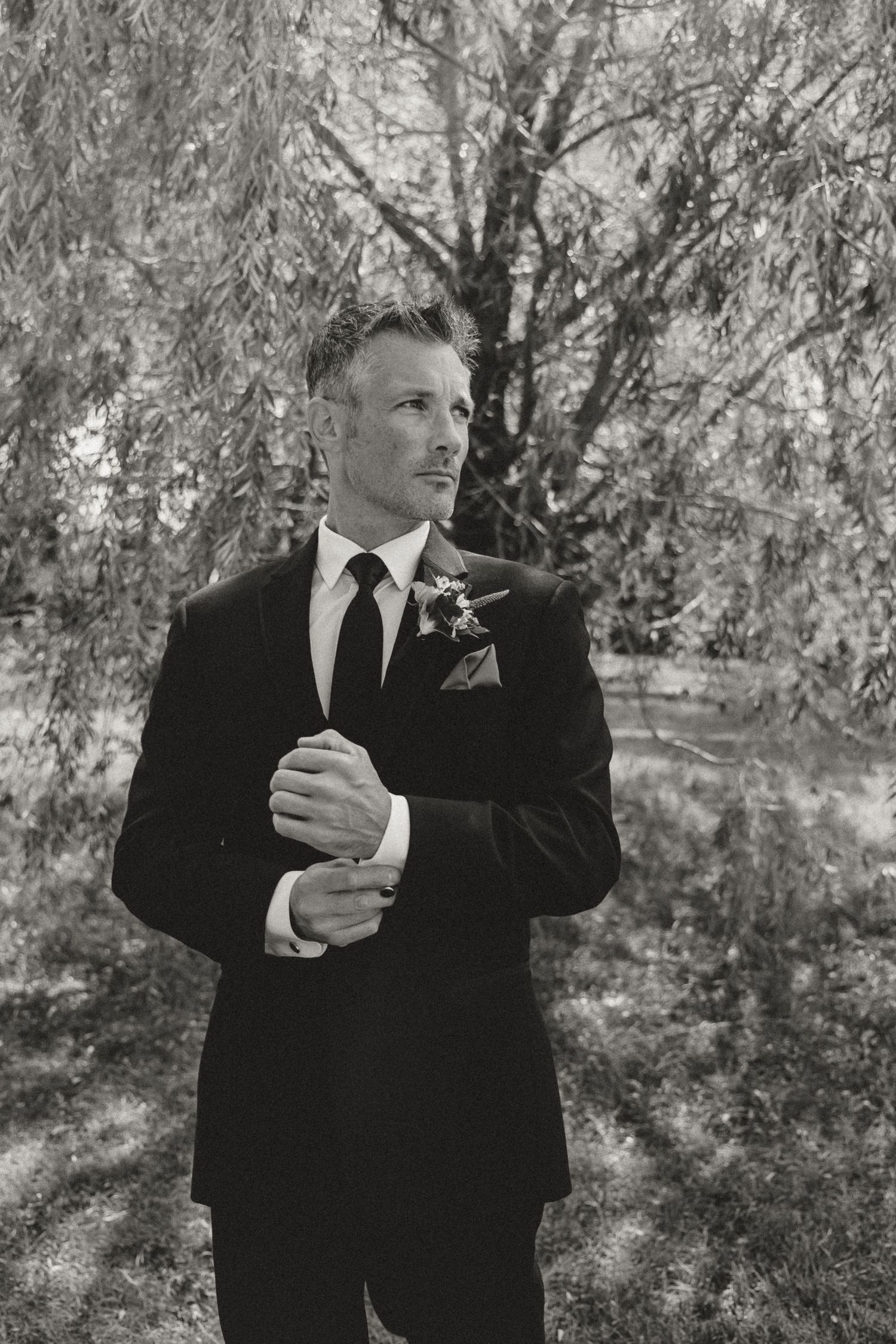 Black and white photo of a groom in a dark suit adjusting cufflinks outdoors under a willow tree.