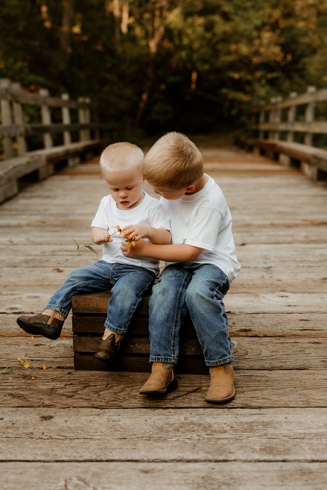 Two young brothers in white shirts and jeans sit together on a wooden bridge, sharing a tender moment outdoors.