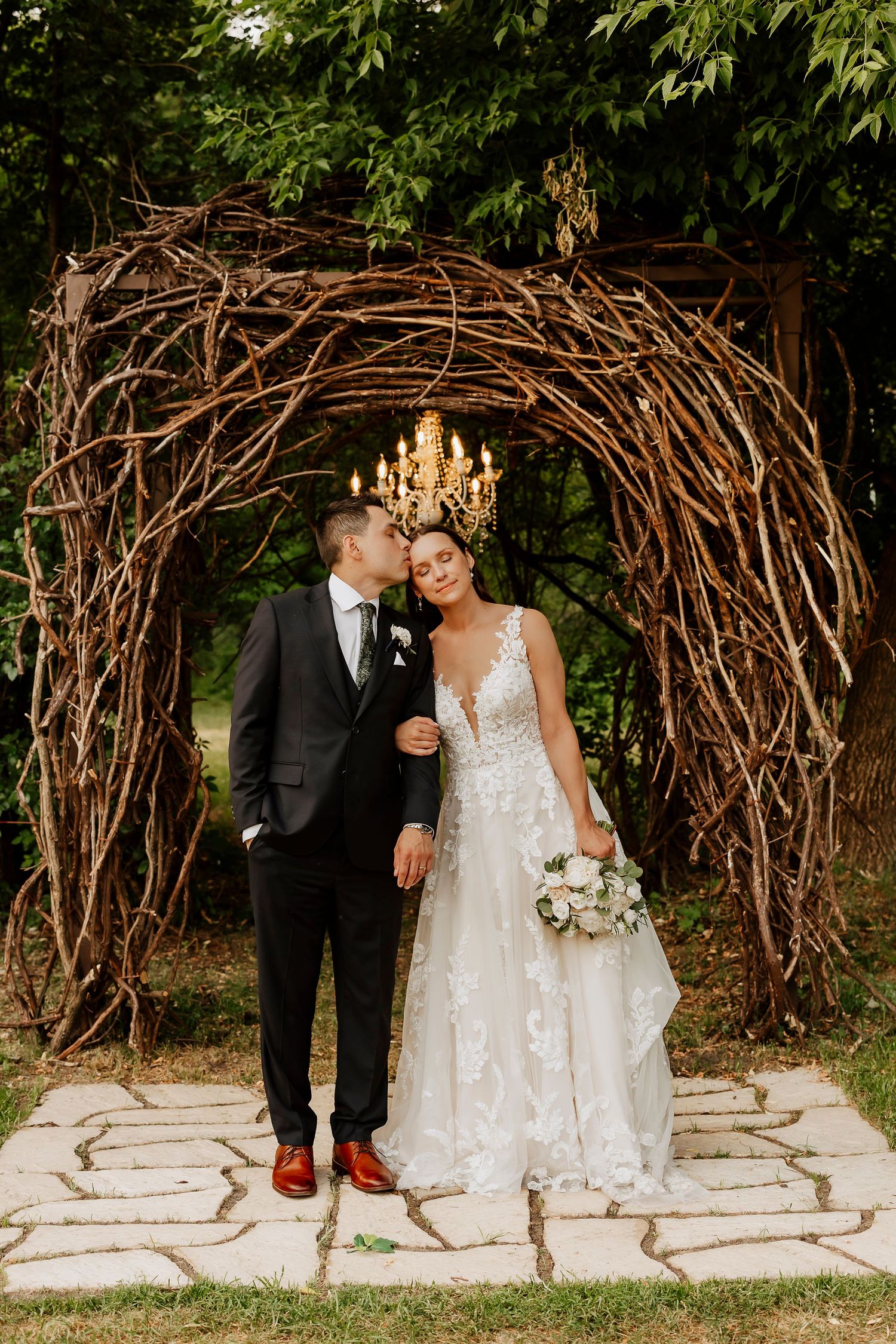 Bride and groom posing under rustic twig arch with chandelier, groom kissing bride's cheek in garden setting.
