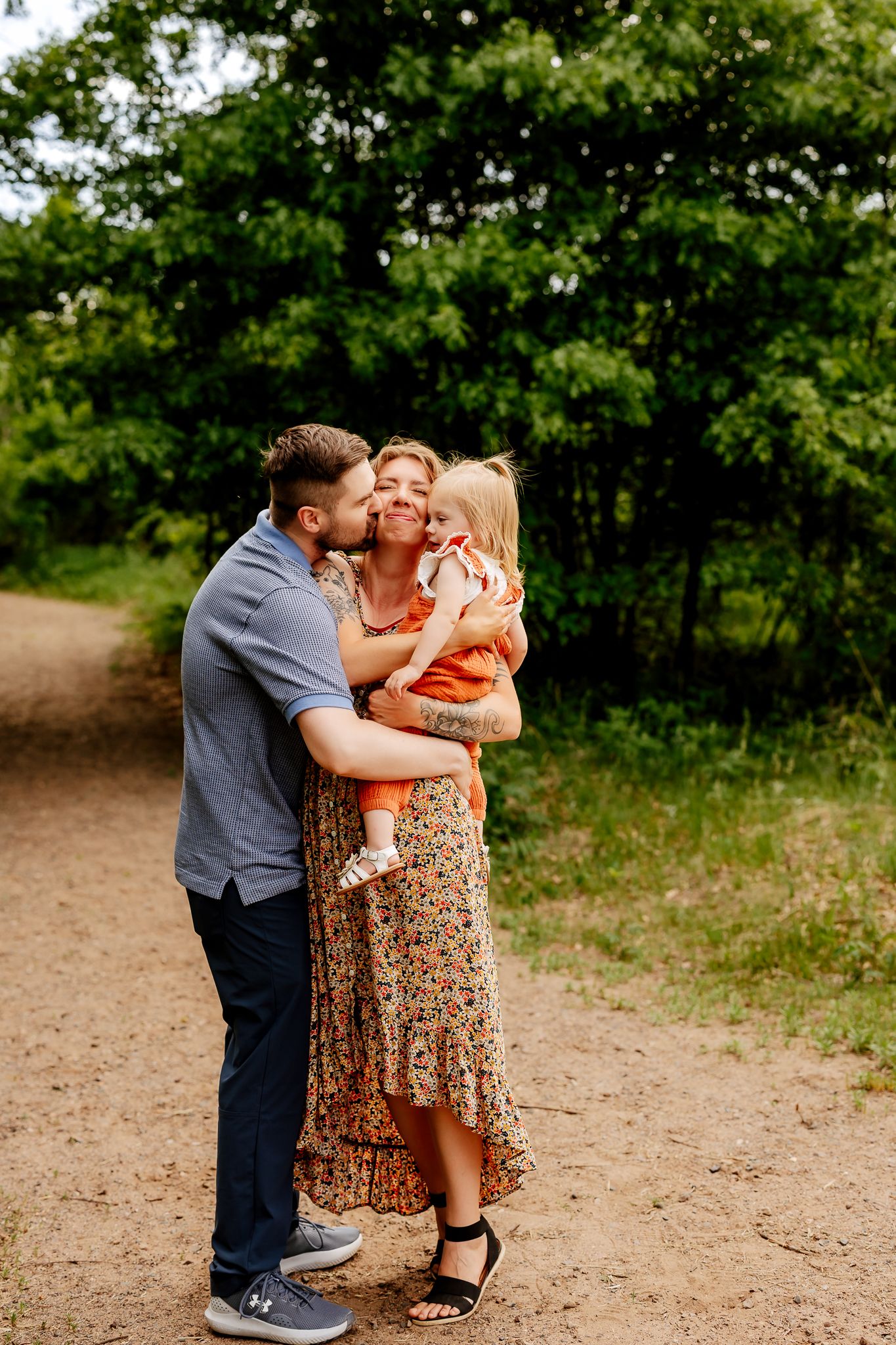 Parents kissing and holding their toddler on a sunlit nature trail.
