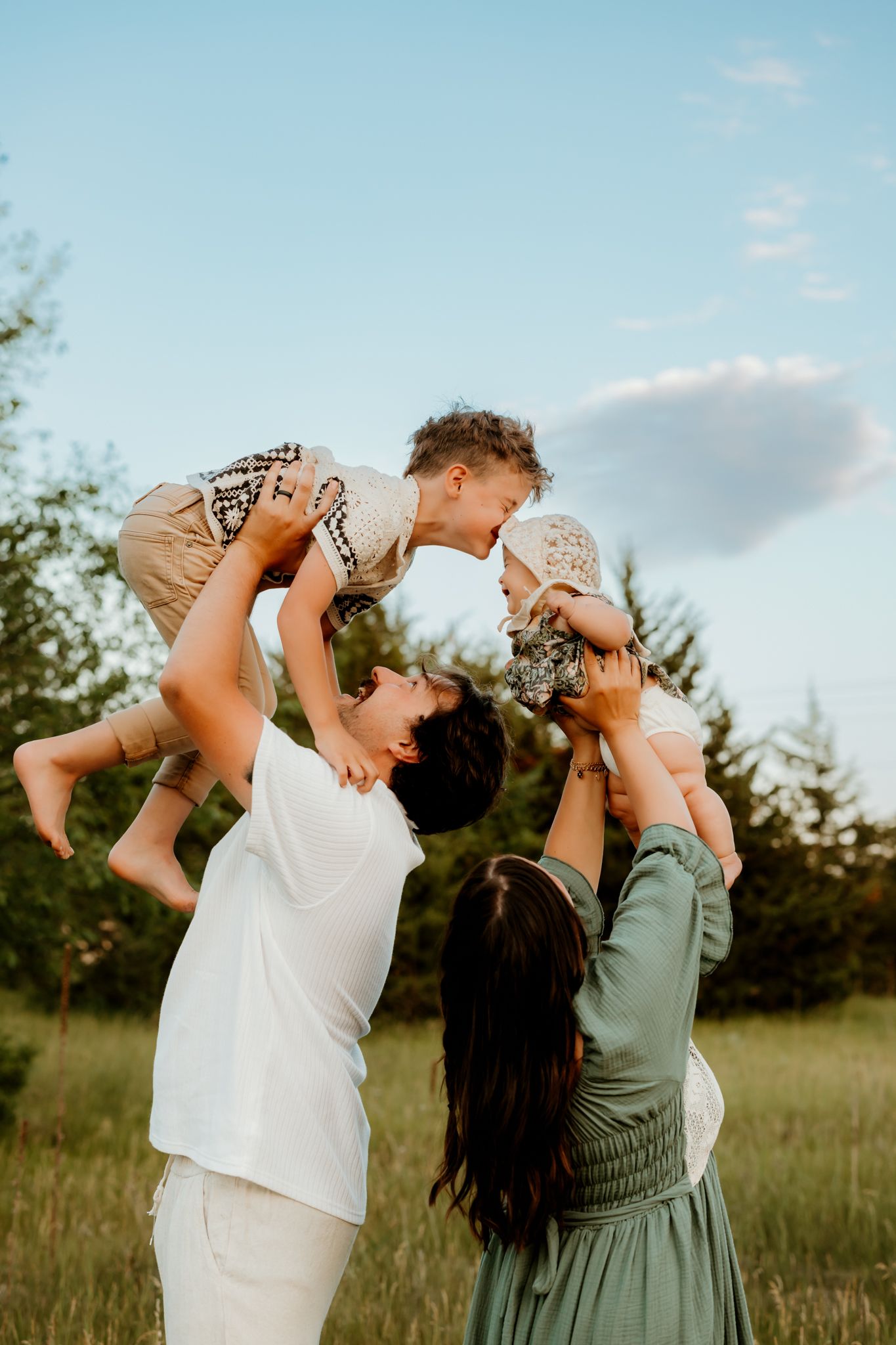 A joyful family outdoors lifting two young children up in the air against a blue sky backdrop.