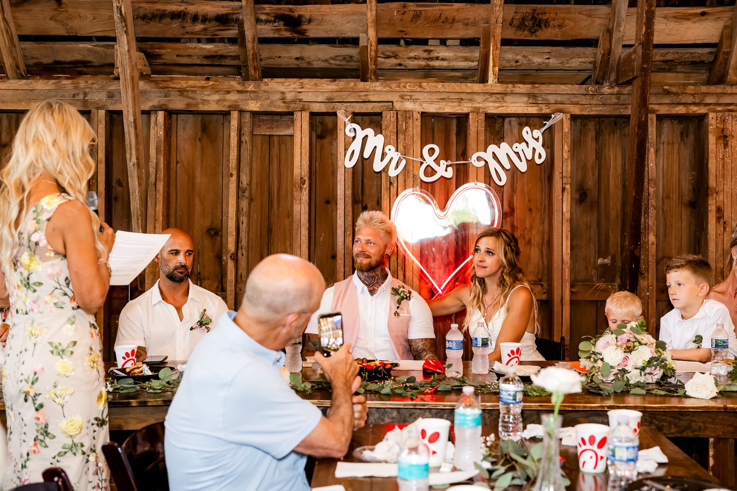 Bride and groom seated at rustic barn wedding reception under Mr. & Mrs. sign with guests celebrating.