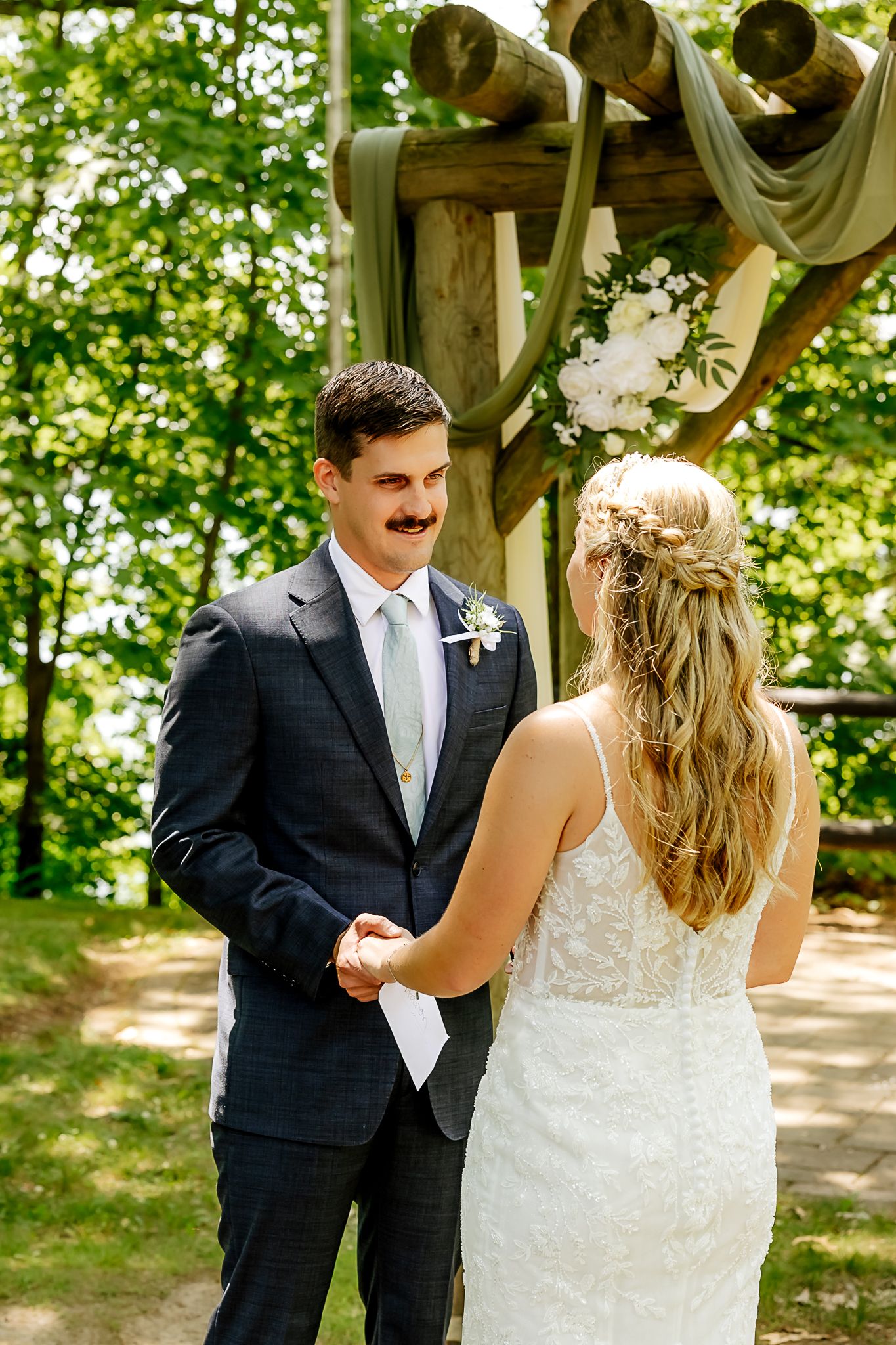 Groom in navy suit holds bride's hands at outdoor wedding ceremony under rustic wooden arch with white flowers.