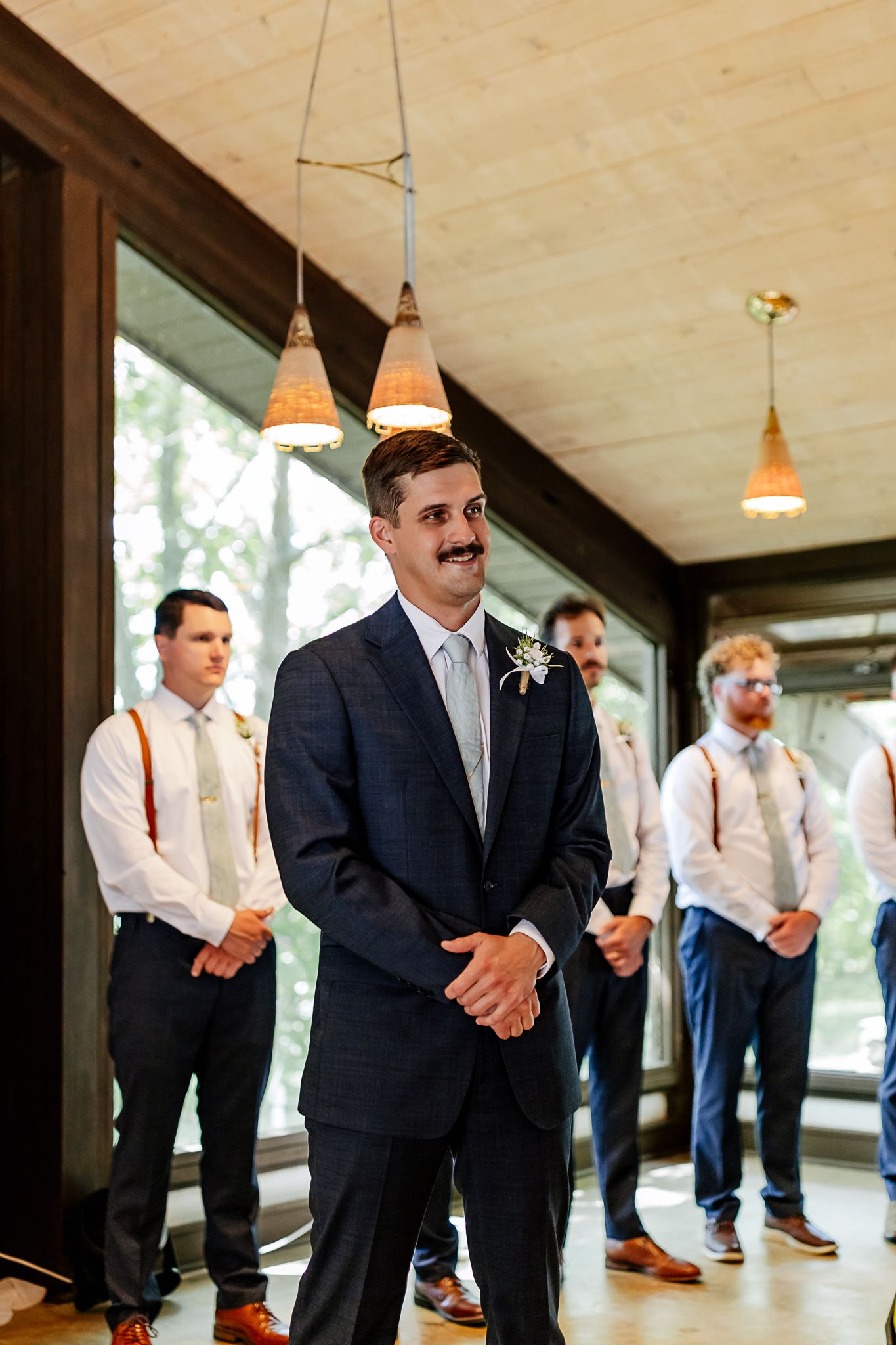 Groom in navy suit with boutonniere smiling at altar, groomsmen in white shirts and suspenders standing behind him.
